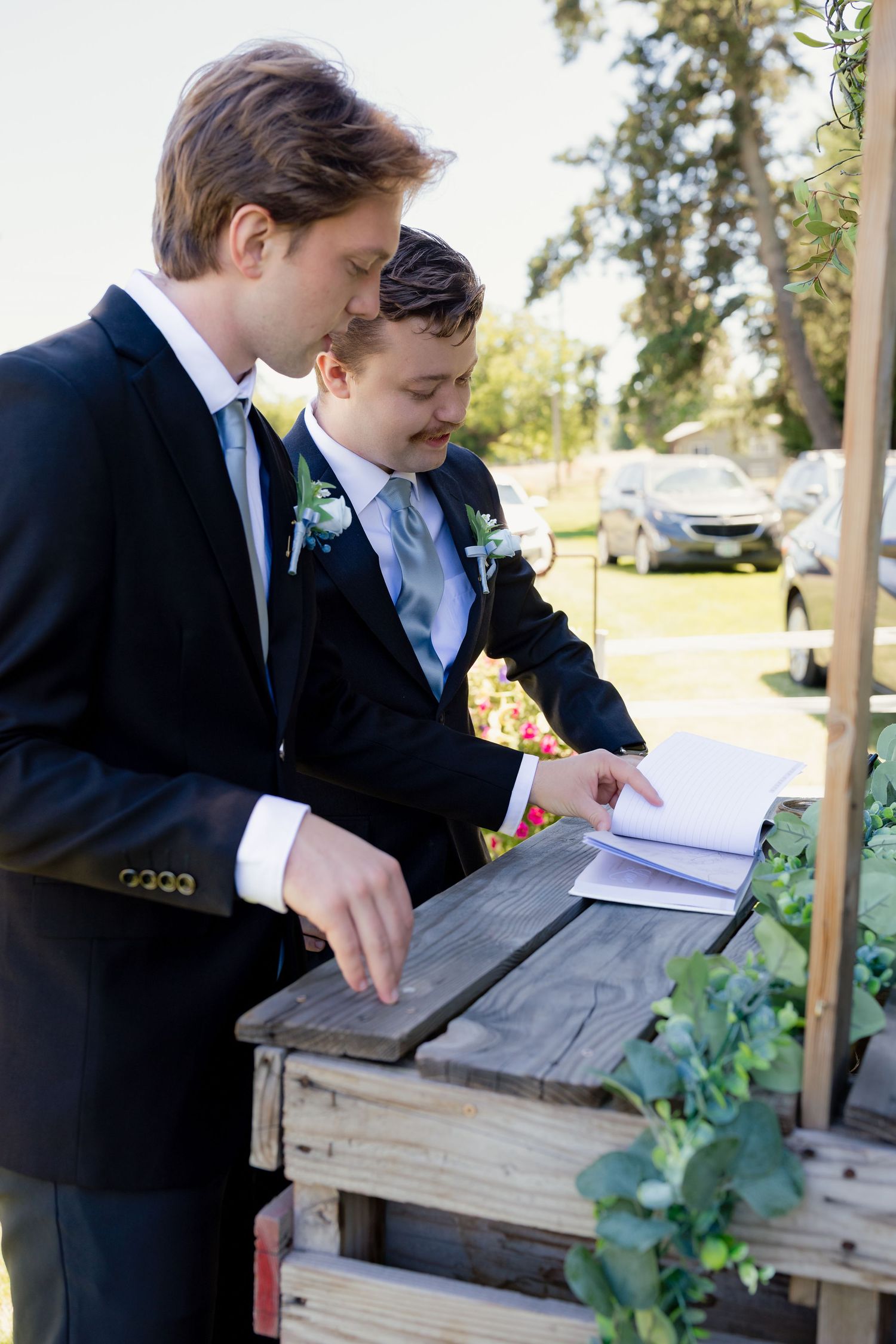 Two groomsmen in black suits review wedding ceremony programs at a rustic wooden stand outdoors.