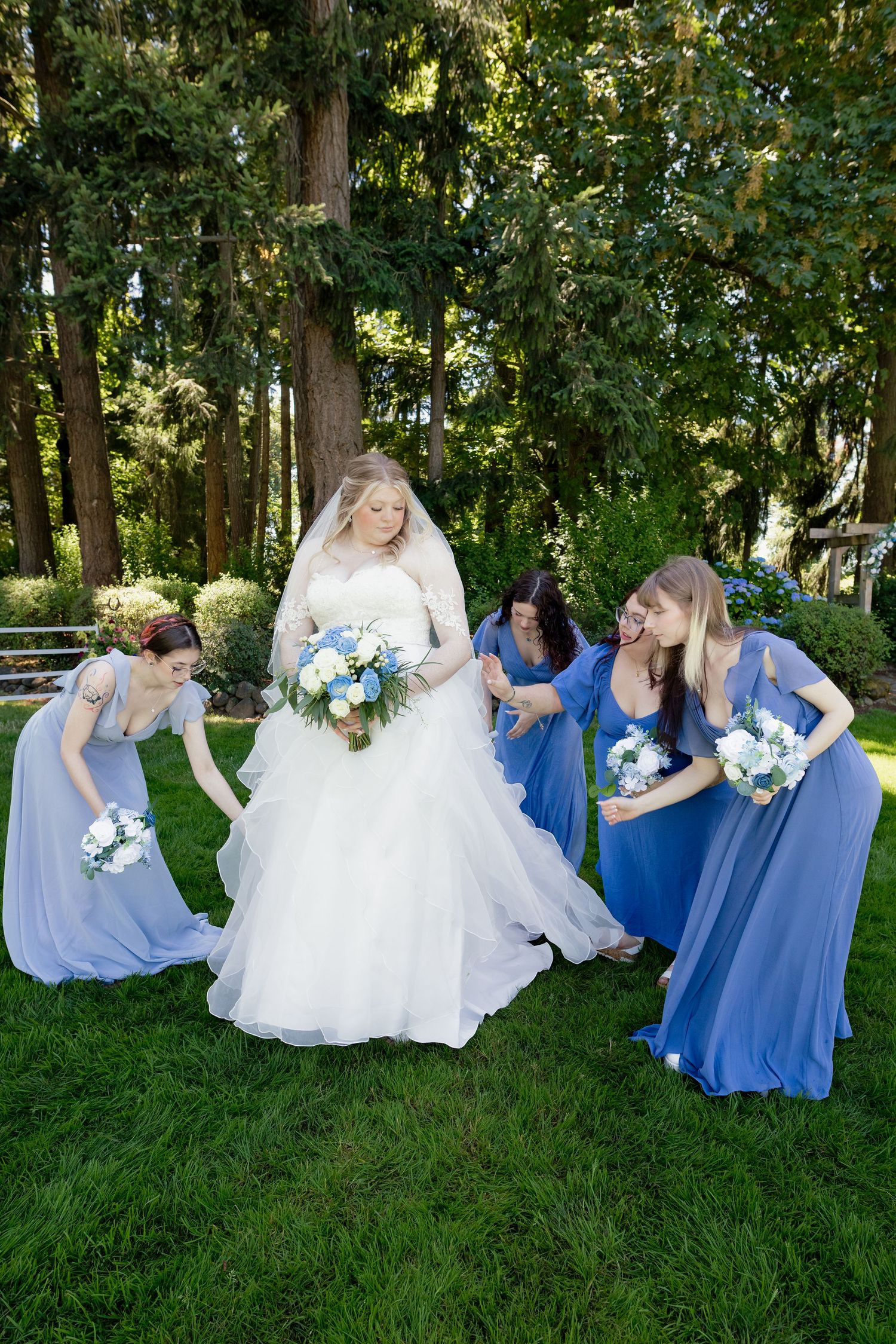 Bride in white gown with bridesmaids in blue dresses holding bouquets in a lush garden setting.