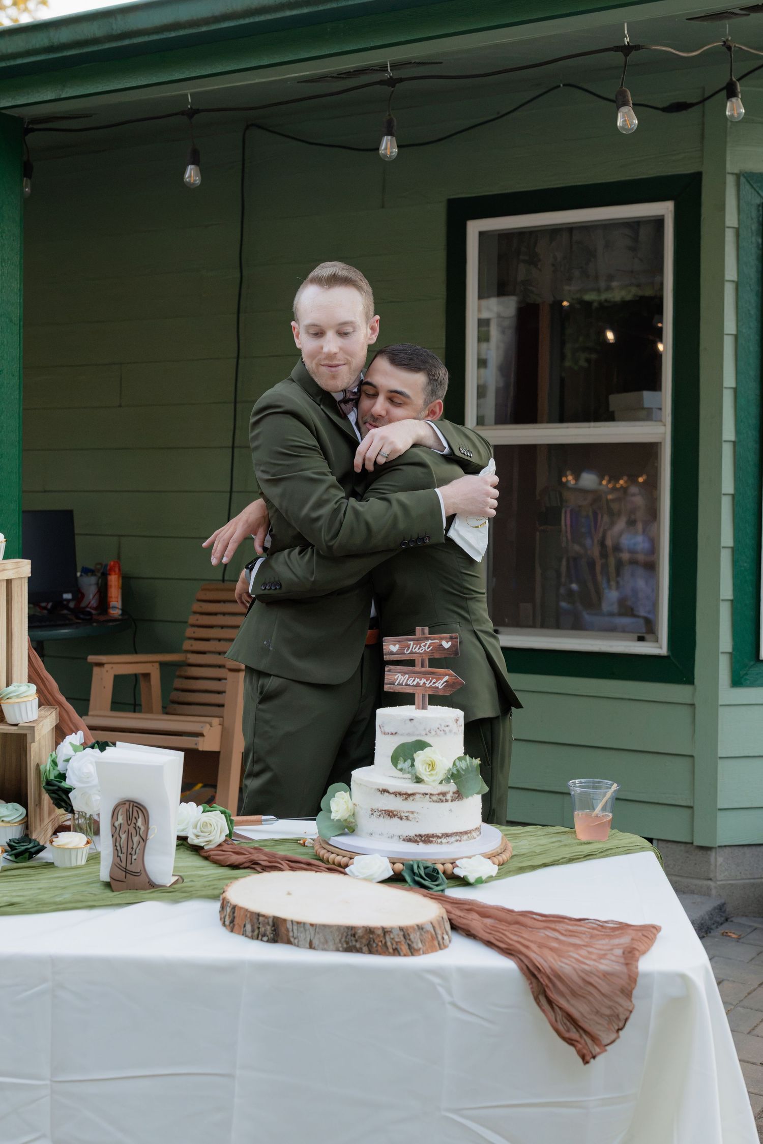 Two grooms in olive green suits share a heartfelt embrace at their wedding cake table outdoors.