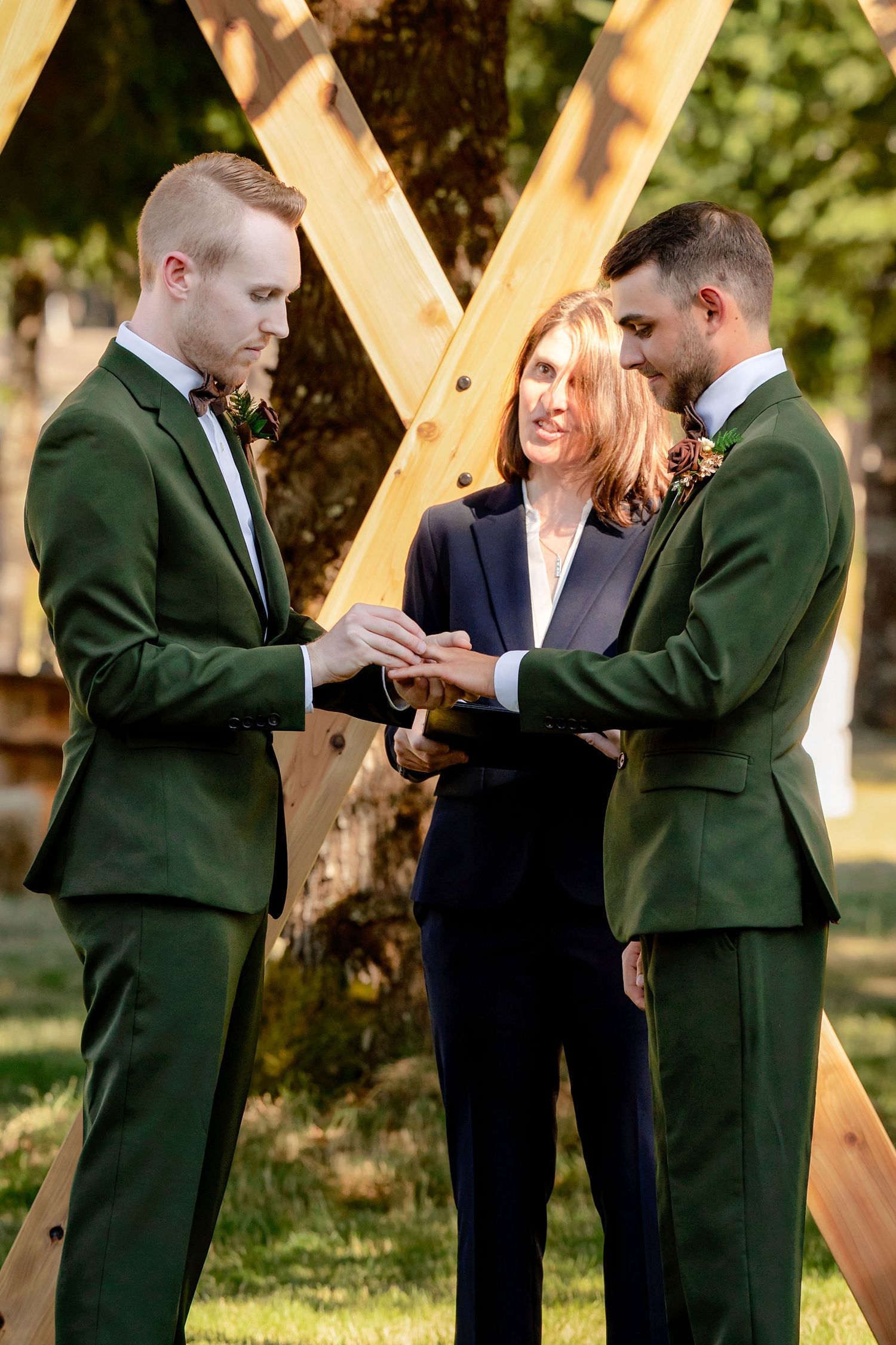 Two grooms in green suits exchanging rings during an outdoor wedding ceremony with wooden arch.