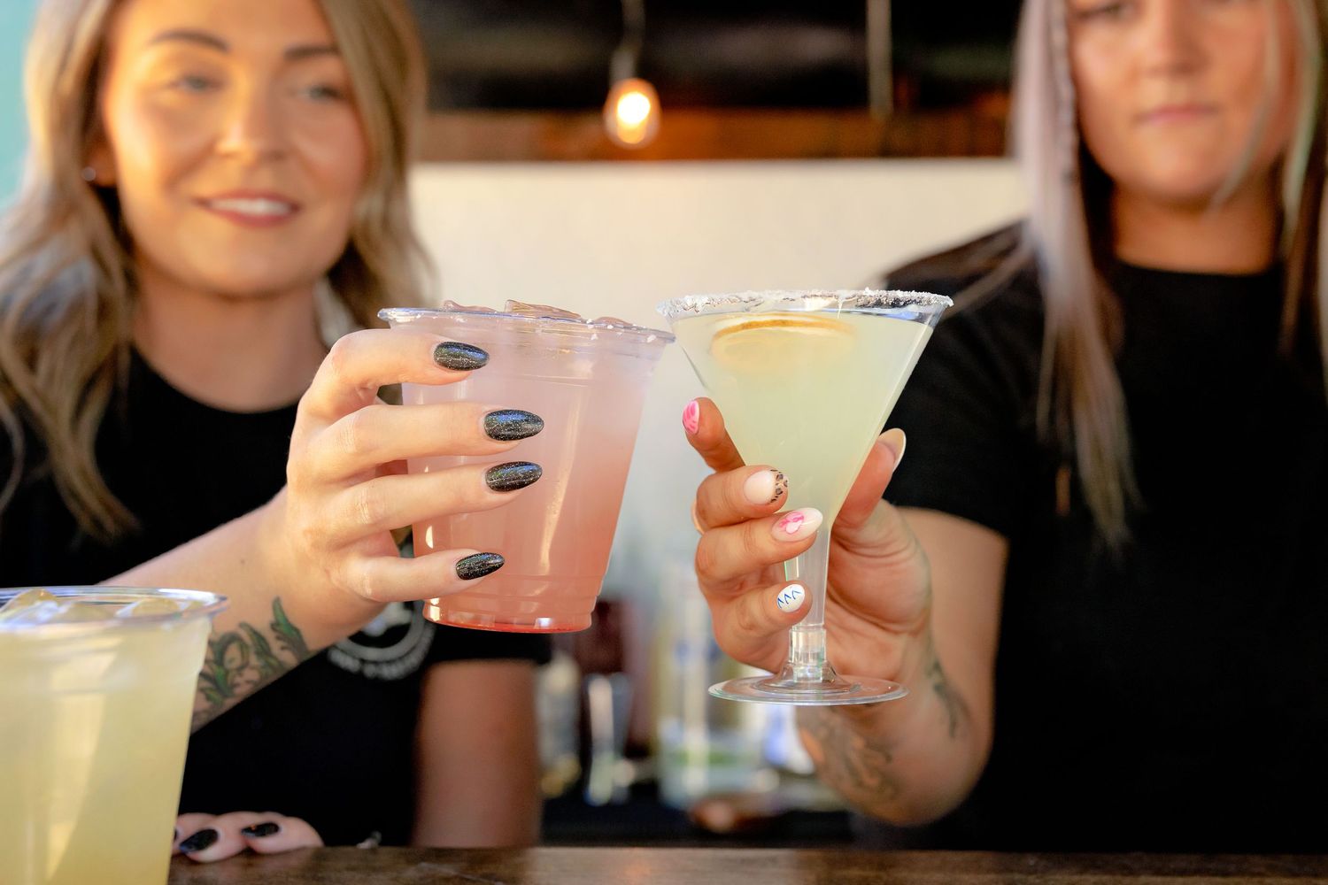 Two women toasting with colorful cocktails, one pink and one green margarita, at a bar.
