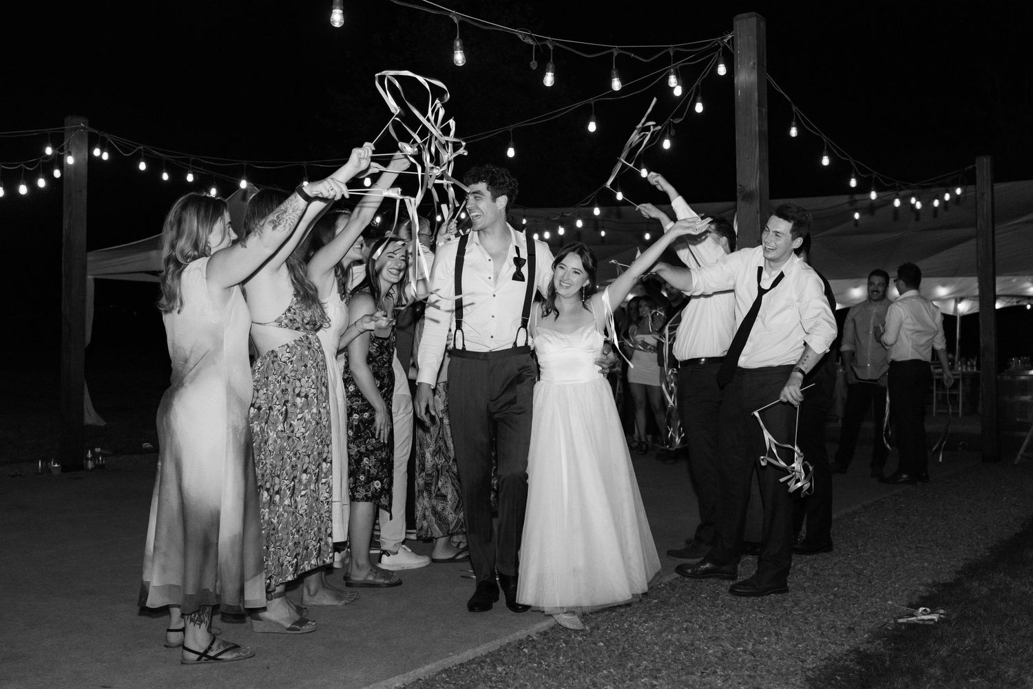 Black and white photo of bride and groom celebrating with guests under string lights at outdoor wedding reception.