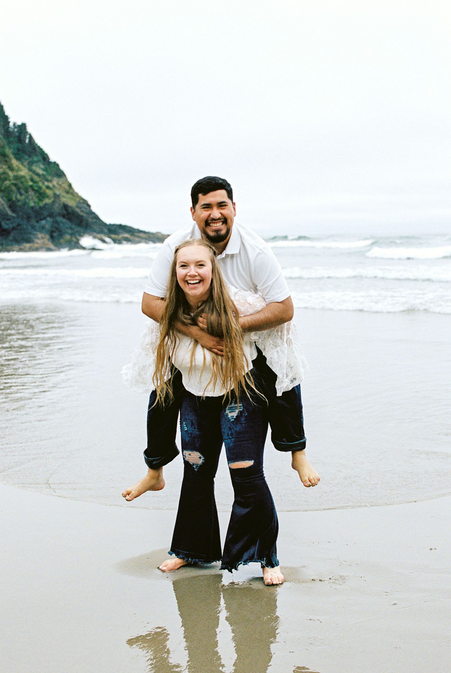 Smiling couple posing on a sandy beach, man lifting woman in white top and black flare jeans with ocean waves behind.