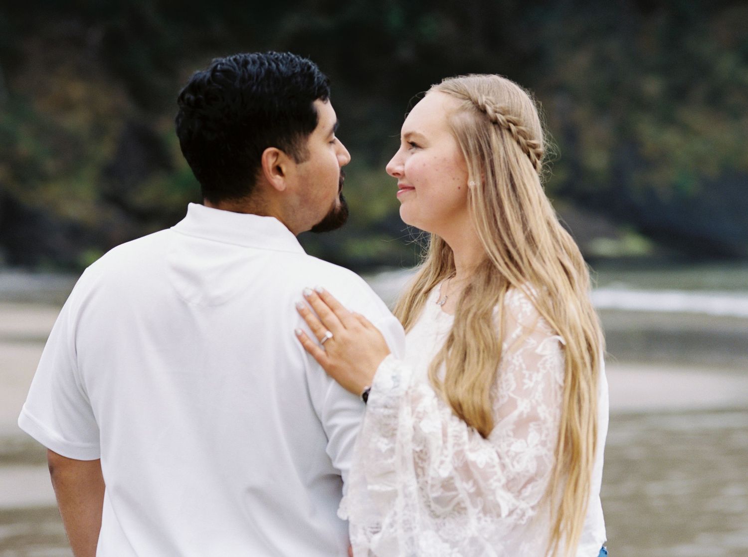 Young couple sharing a romantic moment on a beach, woman in lace dress with braided hair smiling at her partner.
