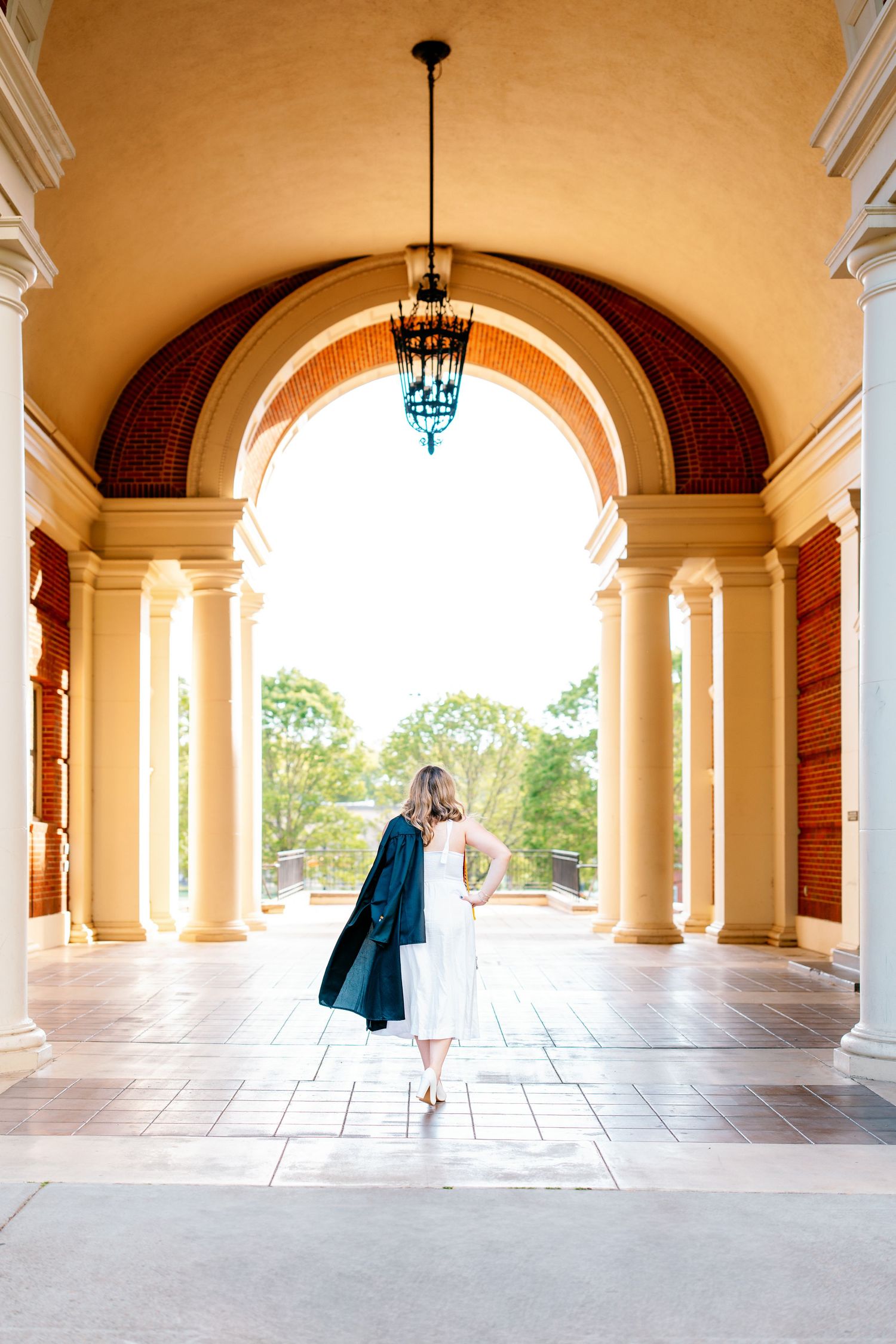 Graduate in white dress and cap walks through elegant arched colonnade with chandelier and warm yellow walls.