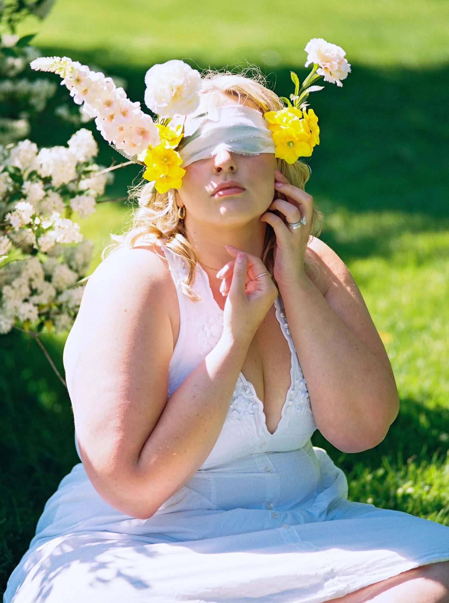 Blindfolded woman in white dress wearing floral crown with yellow and pink flowers, seated outdoors.