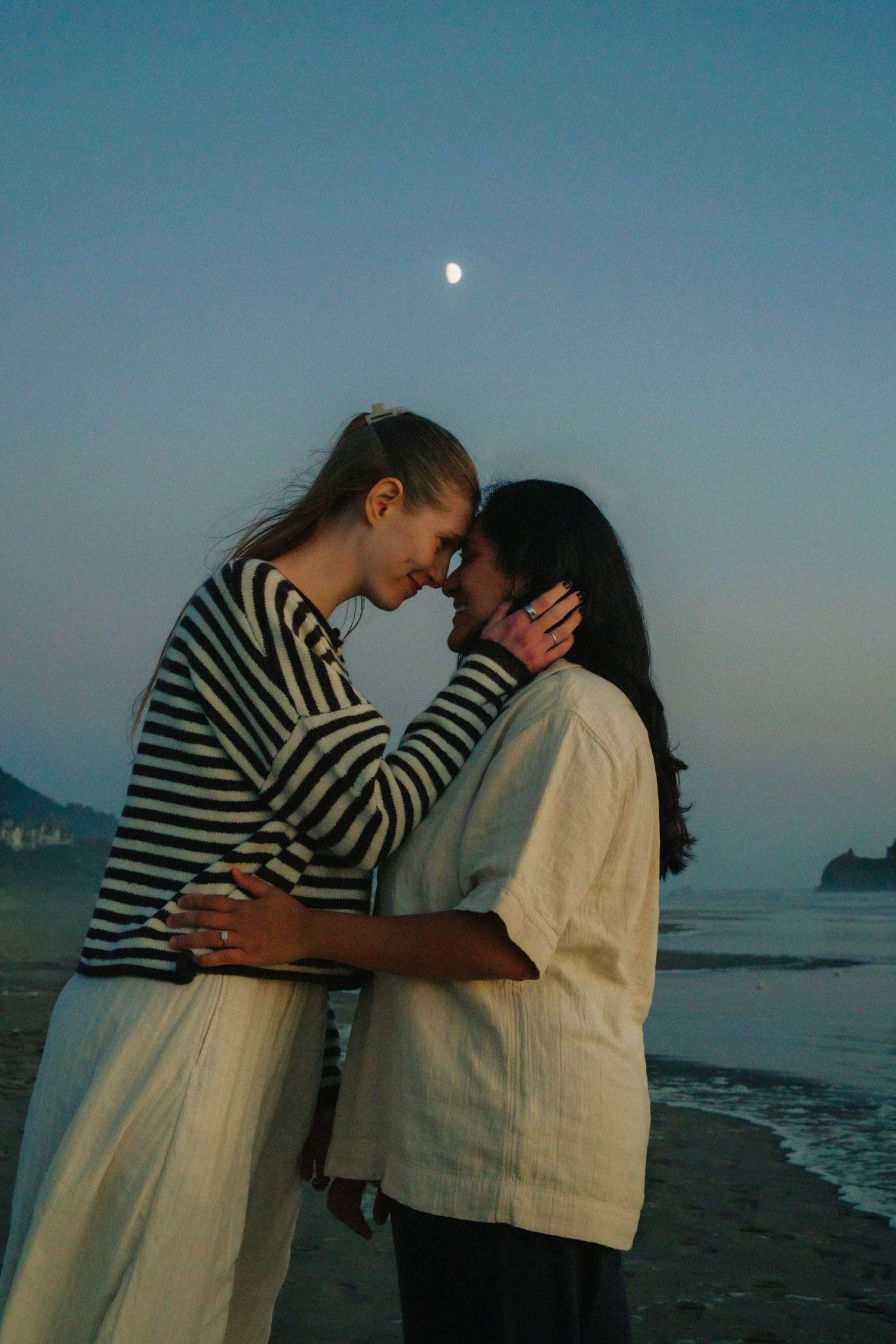 Romantic couple embracing on a beach at dusk with the moon visible in the blue evening sky.
