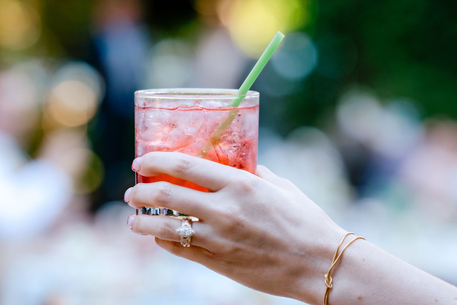 A hand with a gold bracelet and ring holds a pink cocktail with a green straw outdoors.