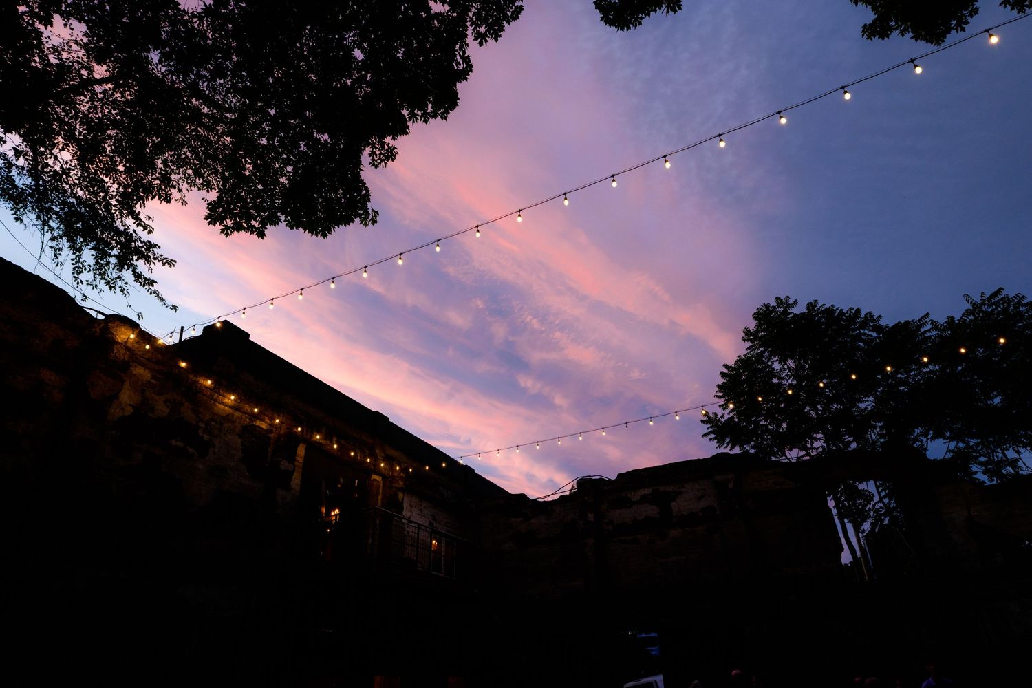 String lights hang against a vibrant pink and blue sunset sky, framed by dark trees and a silhouetted building.