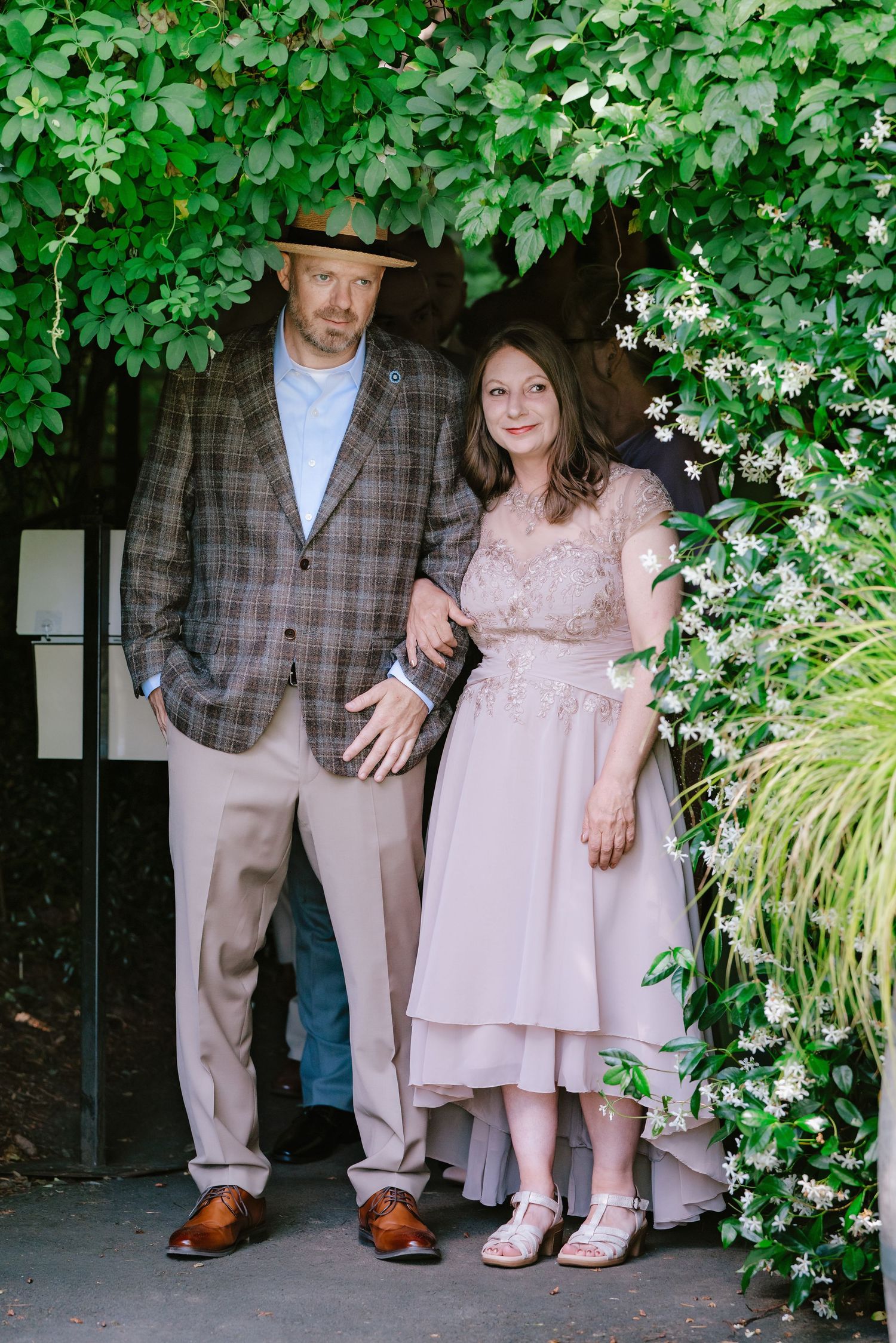 Couple posing under lush green archway, man in plaid blazer and straw hat, woman in blush pink dress.
