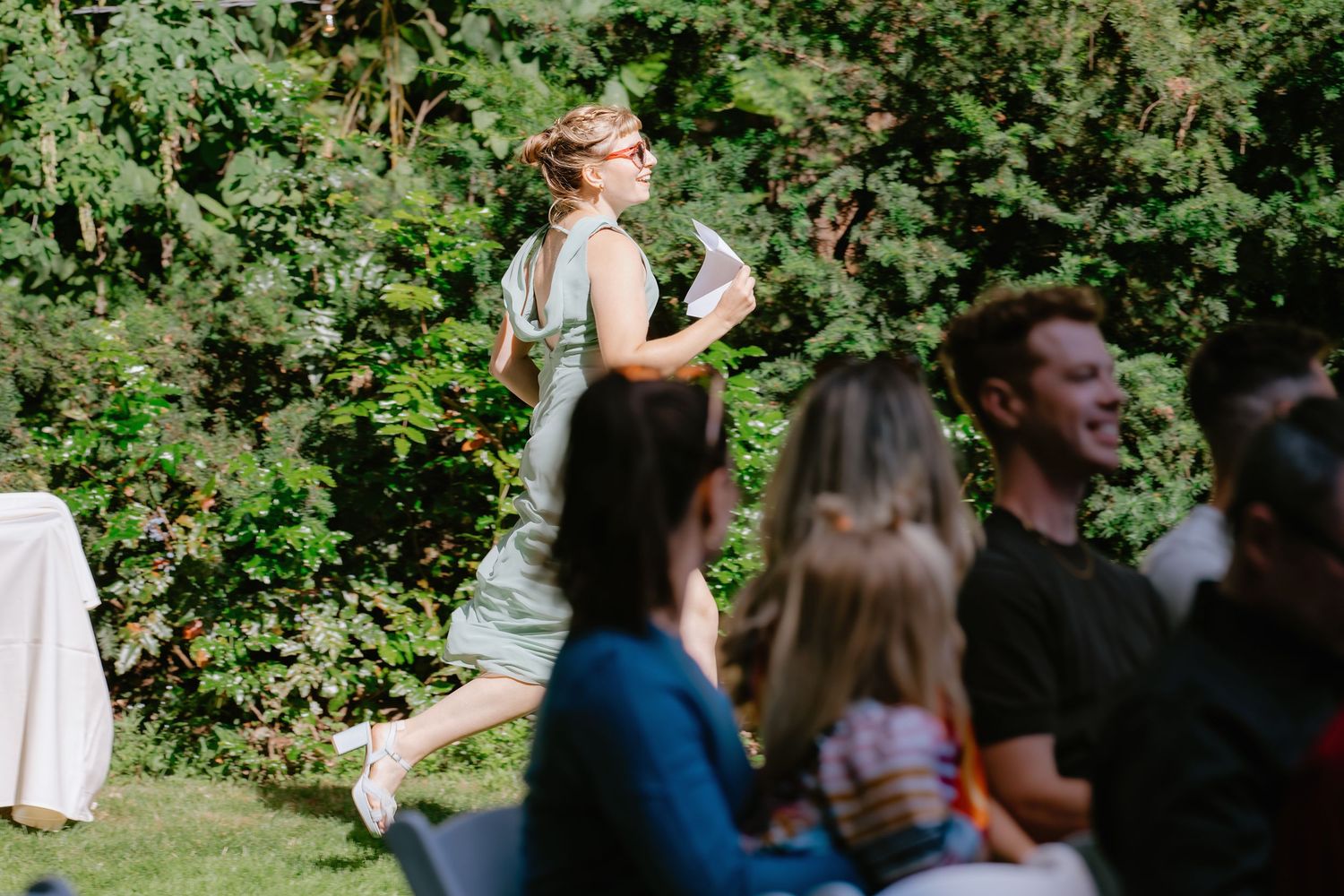 Woman in sage green dress walks briskly past seated wedding guests at outdoor ceremony.