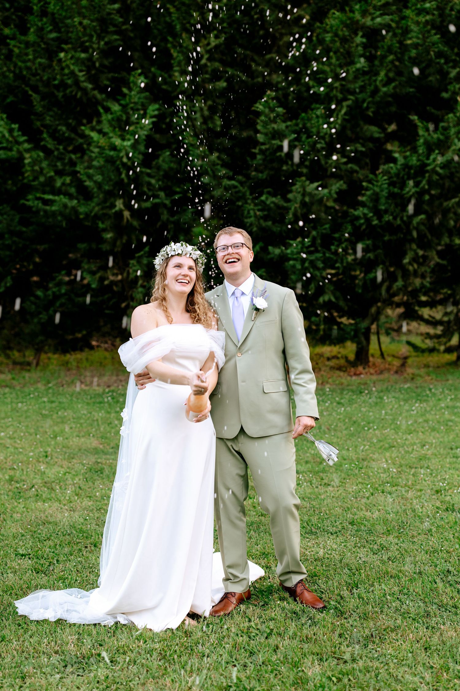 Laughing bride and groom celebrate outdoors as champagne sprays from bottle on green lawn.