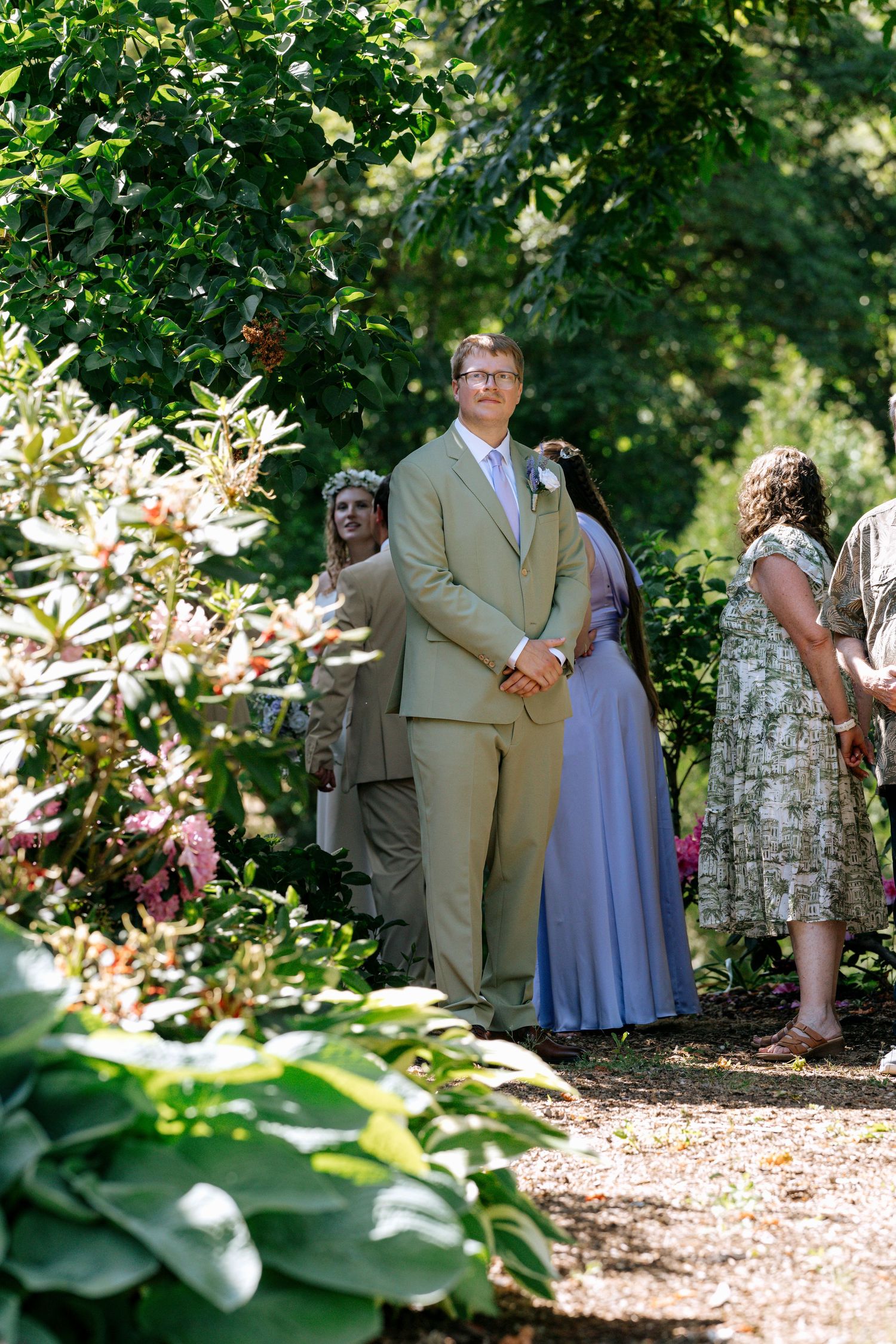 Groom in tan suit stands at outdoor wedding ceremony aisle surrounded by lush green garden foliage.