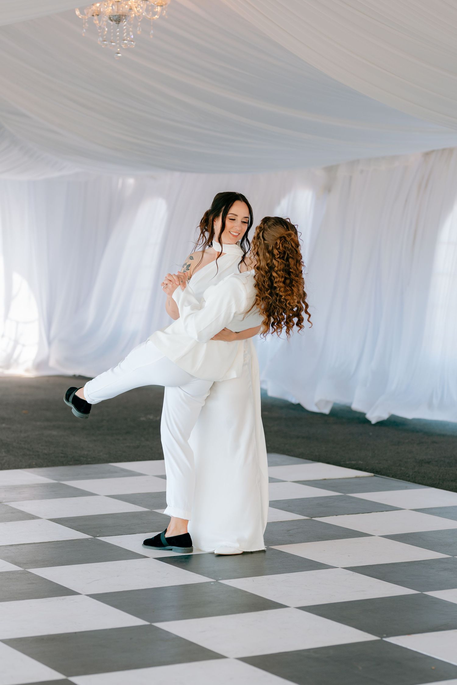 Two brides in white outfits share a joyful first dance on a checkered floor under an elegant draped tent.