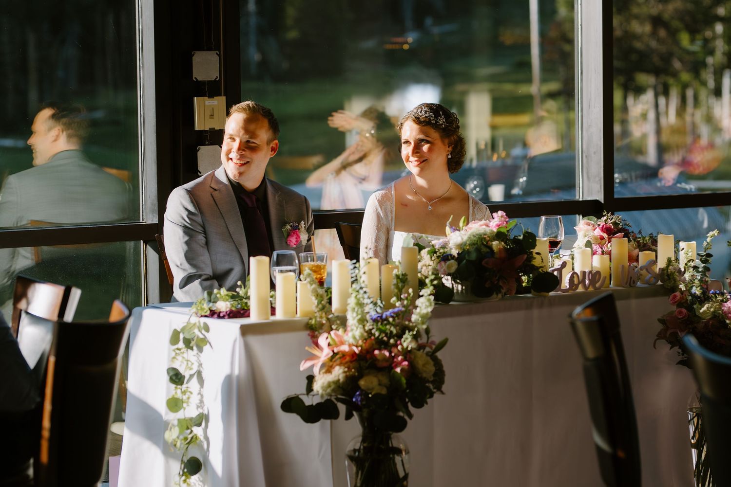 Smiling bride and groom seated together at sweetheart table during wedding reception near large windows.