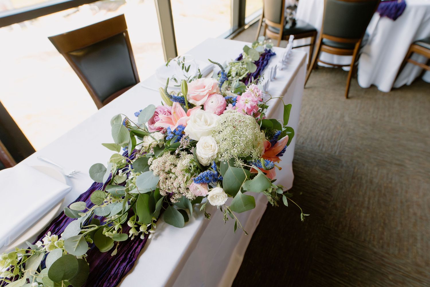 Elegant wedding head table with lush floral centerpiece featuring pink roses, white blooms, and eucalyptus greenery.