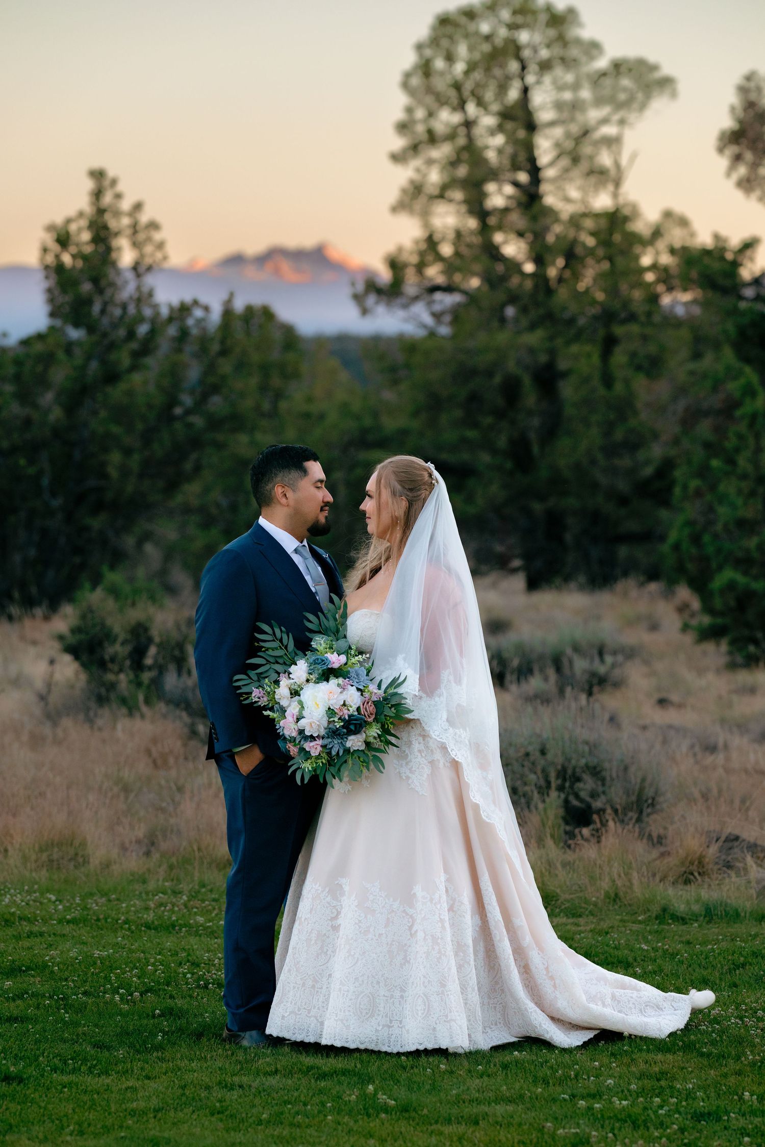 Bride and groom share a romantic moment at sunset, with mountains and trees in the background, as she holds a lush bouquet.