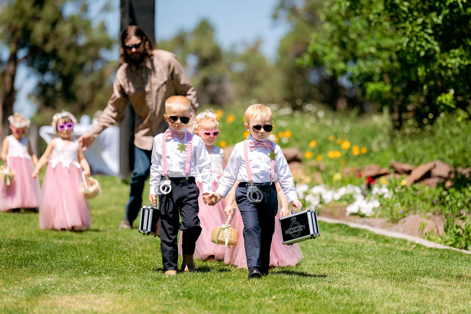 Adorable young ring bearers in suits with sunglasses carry signs down grassy wedding aisle.