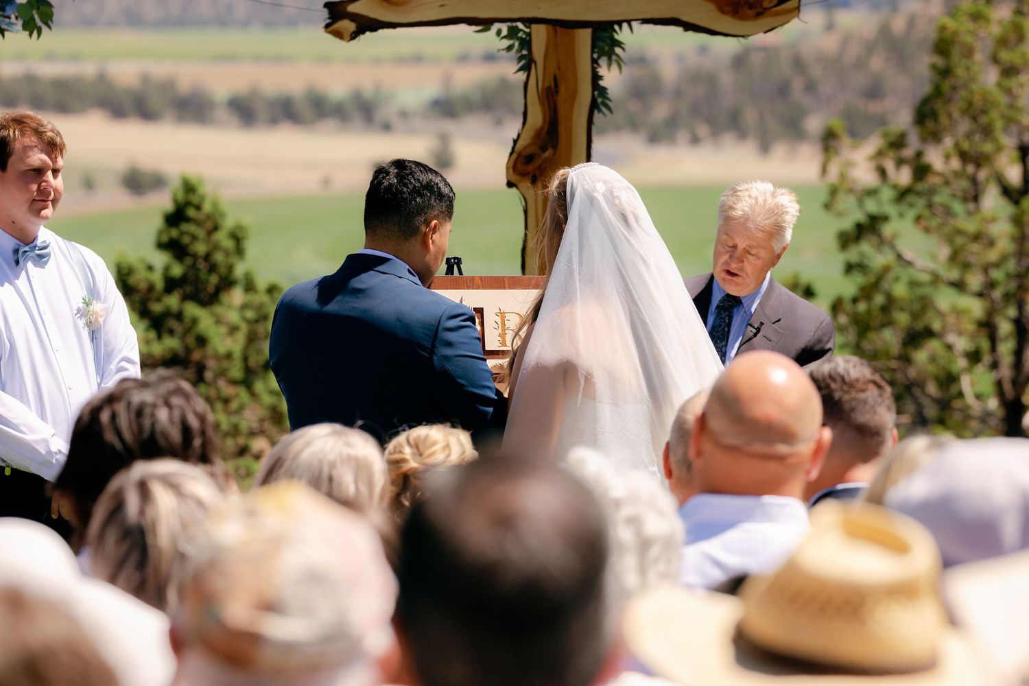 Outdoor wedding ceremony with couple exchanging vows before officiant, scenic green valley backdrop, guests seated in foreground.