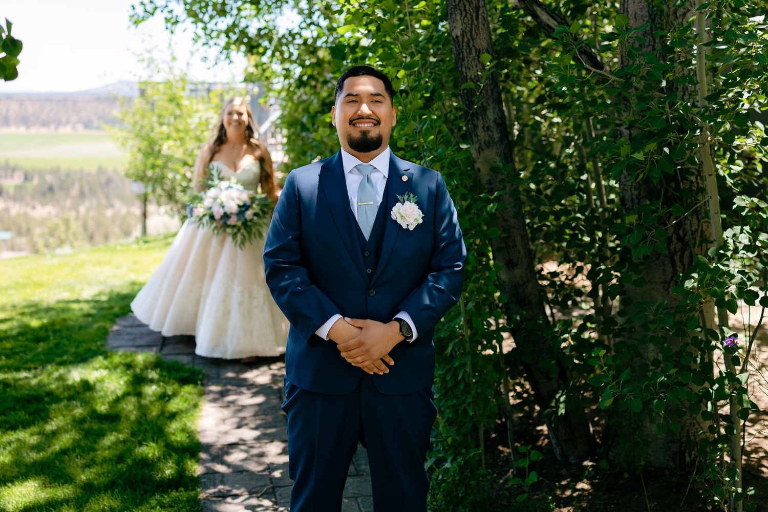 Groom in navy suit awaits bride for first look moment outdoors at wedding ceremony.