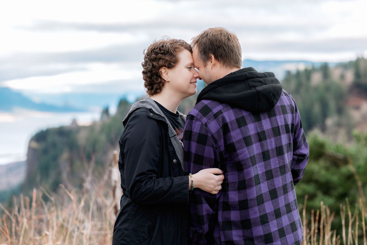 A couple embraces while standing in front of a mountainous landscape on an overcast day.