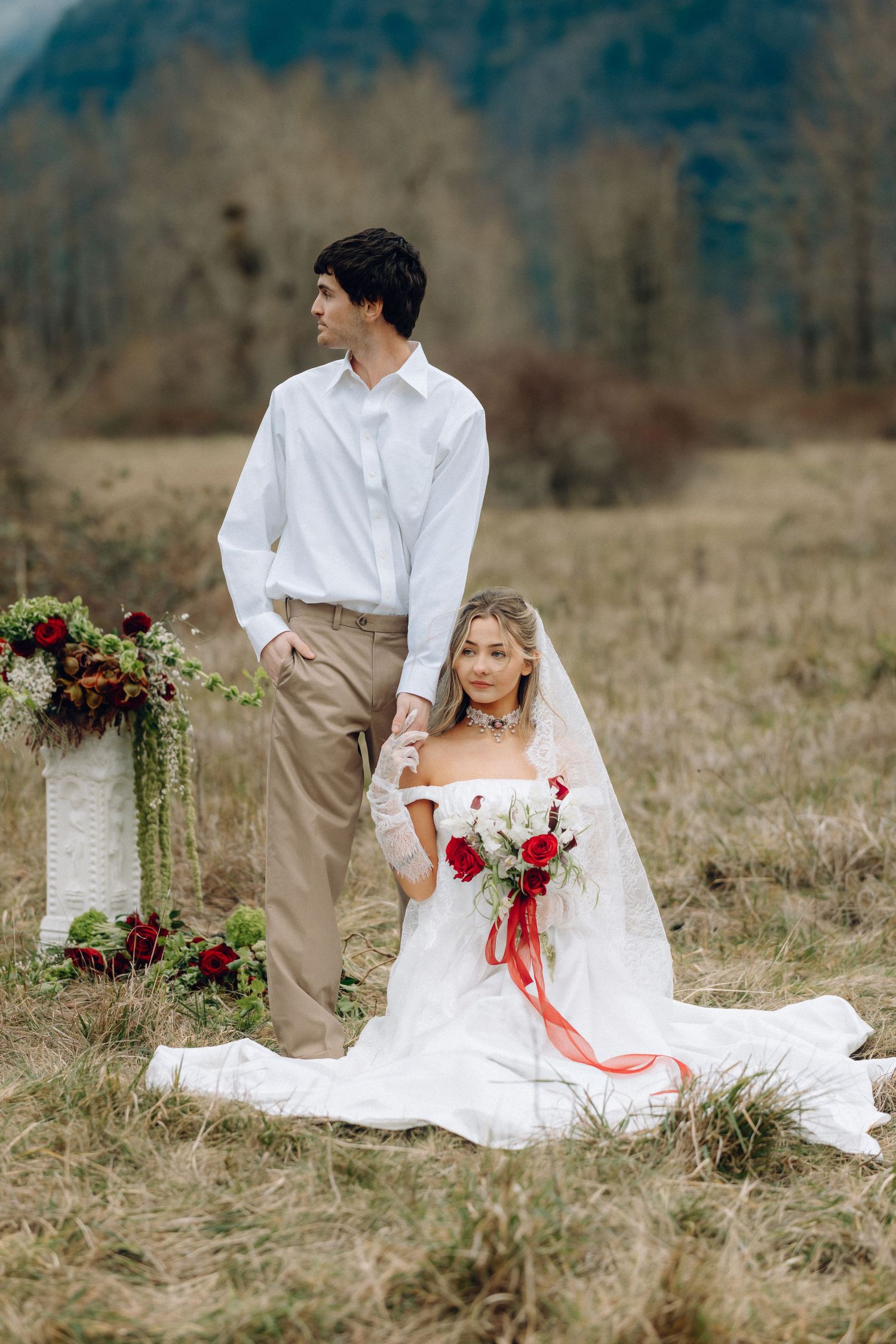A romantic wedding photoshoot in a rustic field with red floral accents and flowing white dress.