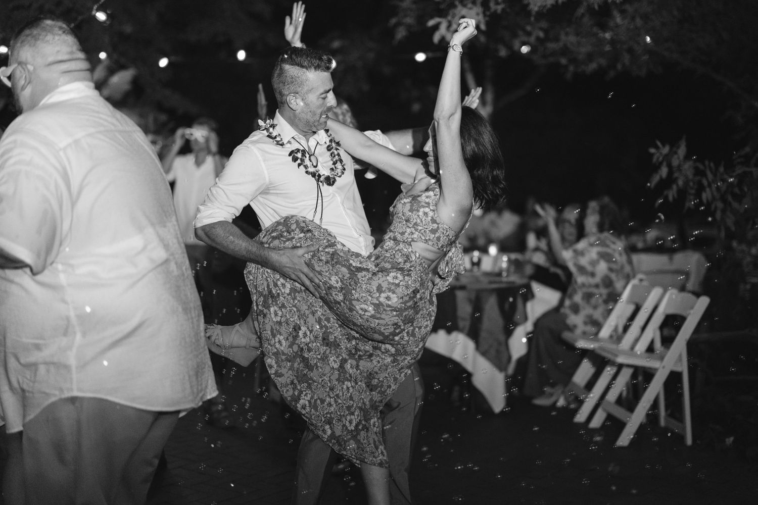 A couple dances energetically at an outdoor reception, with the woman's floral dress twirling in black and white photography.