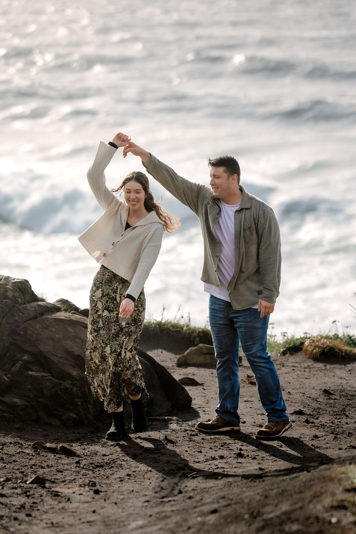 A couple playfully poses together on a rocky beach with waves in the background.