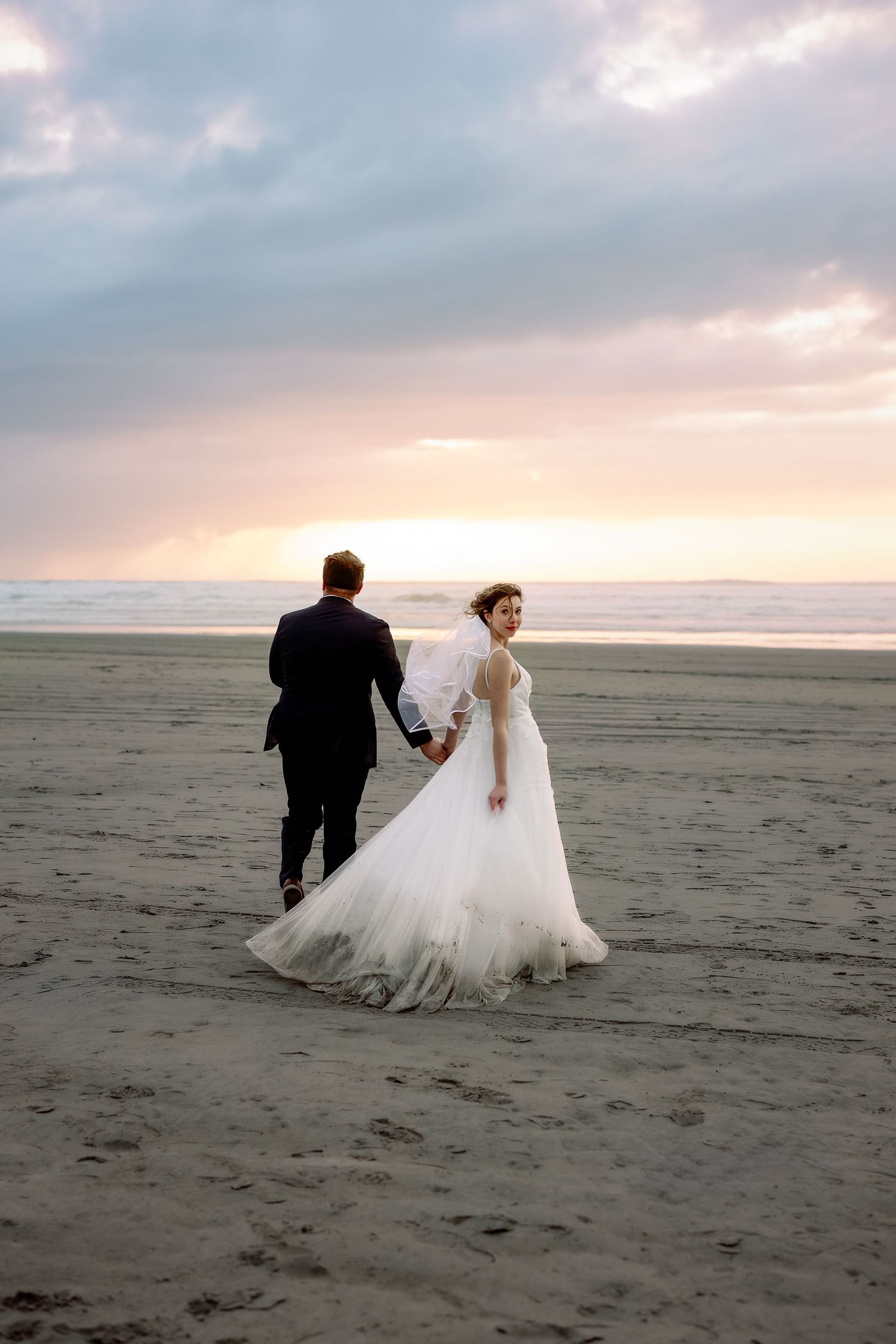 A couple in wedding attire walks along a misty beach at sunset, their silhouettes casting long shadows on the sand.