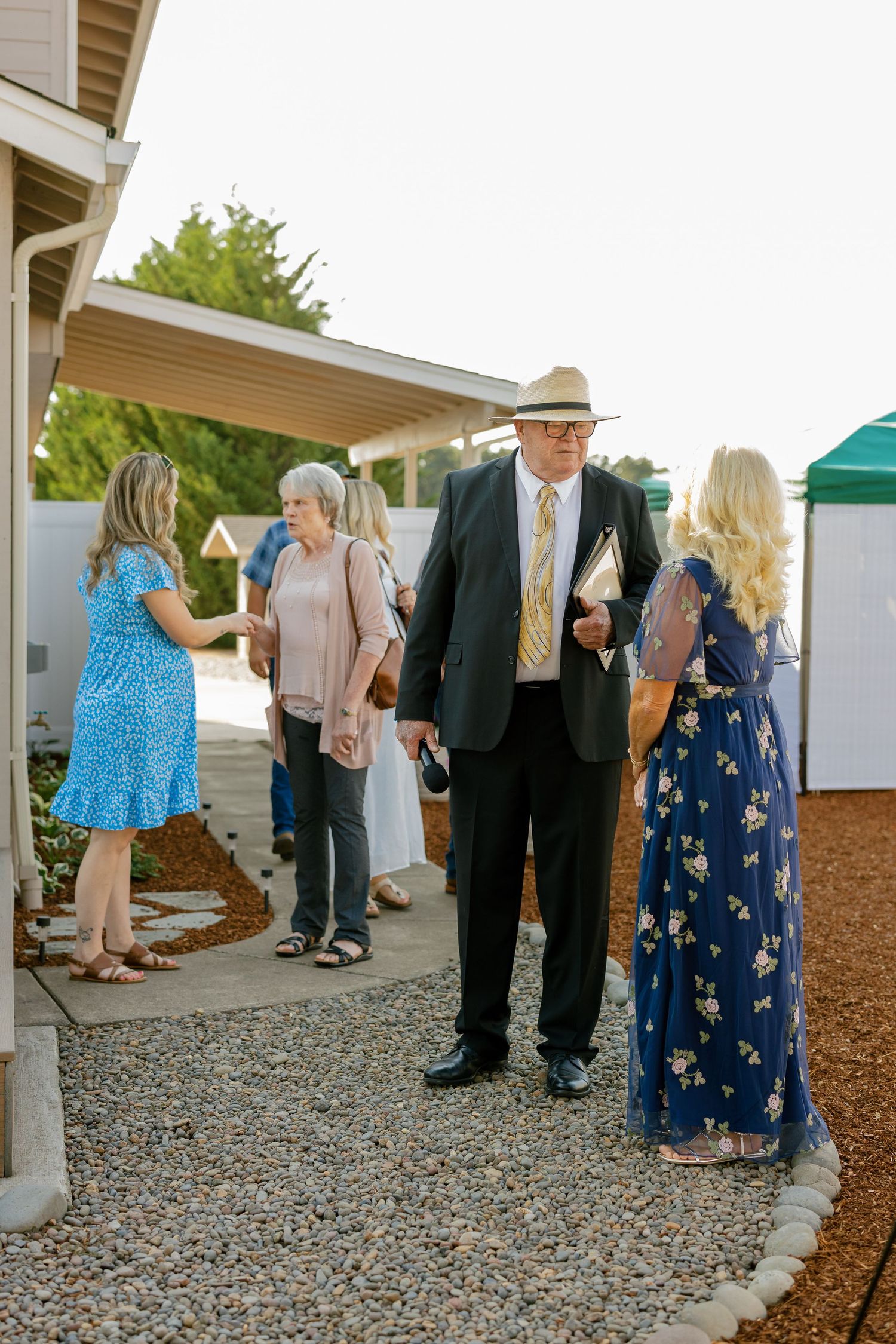 Wedding guests gather on a gravel path outside a covered porch during an outdoor reception.