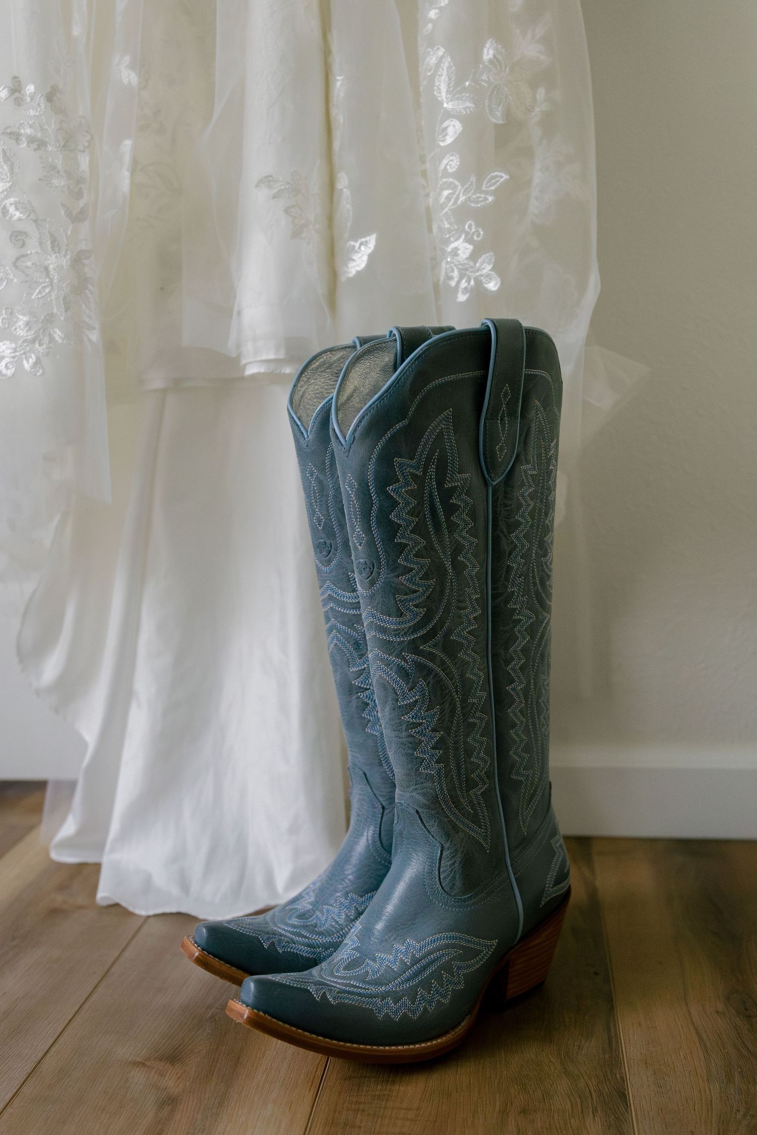 Blue cowboy boots displayed against white lace wedding dress on wooden floor.