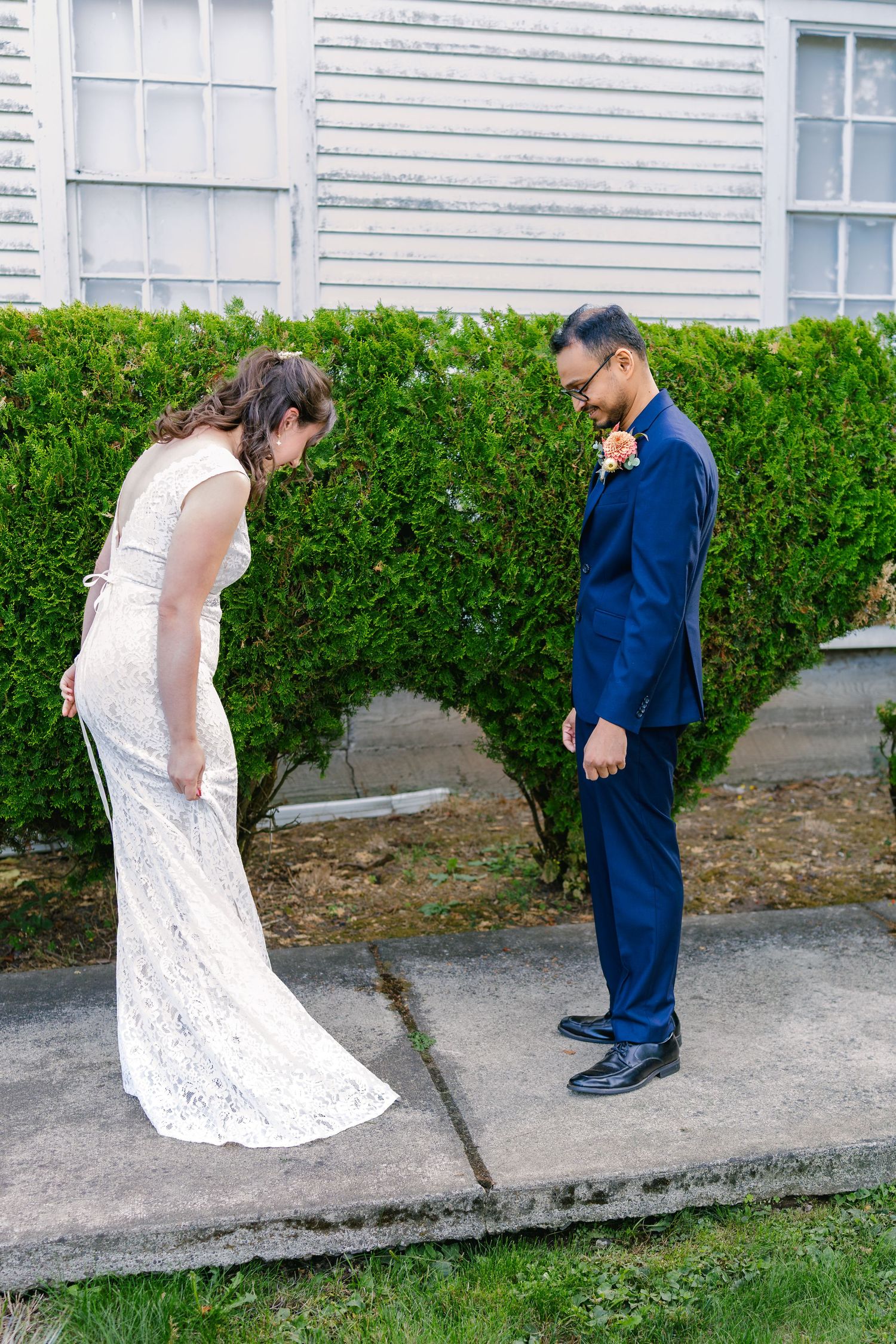Couple in wedding attire walking and interacting along a white clapboard building.
