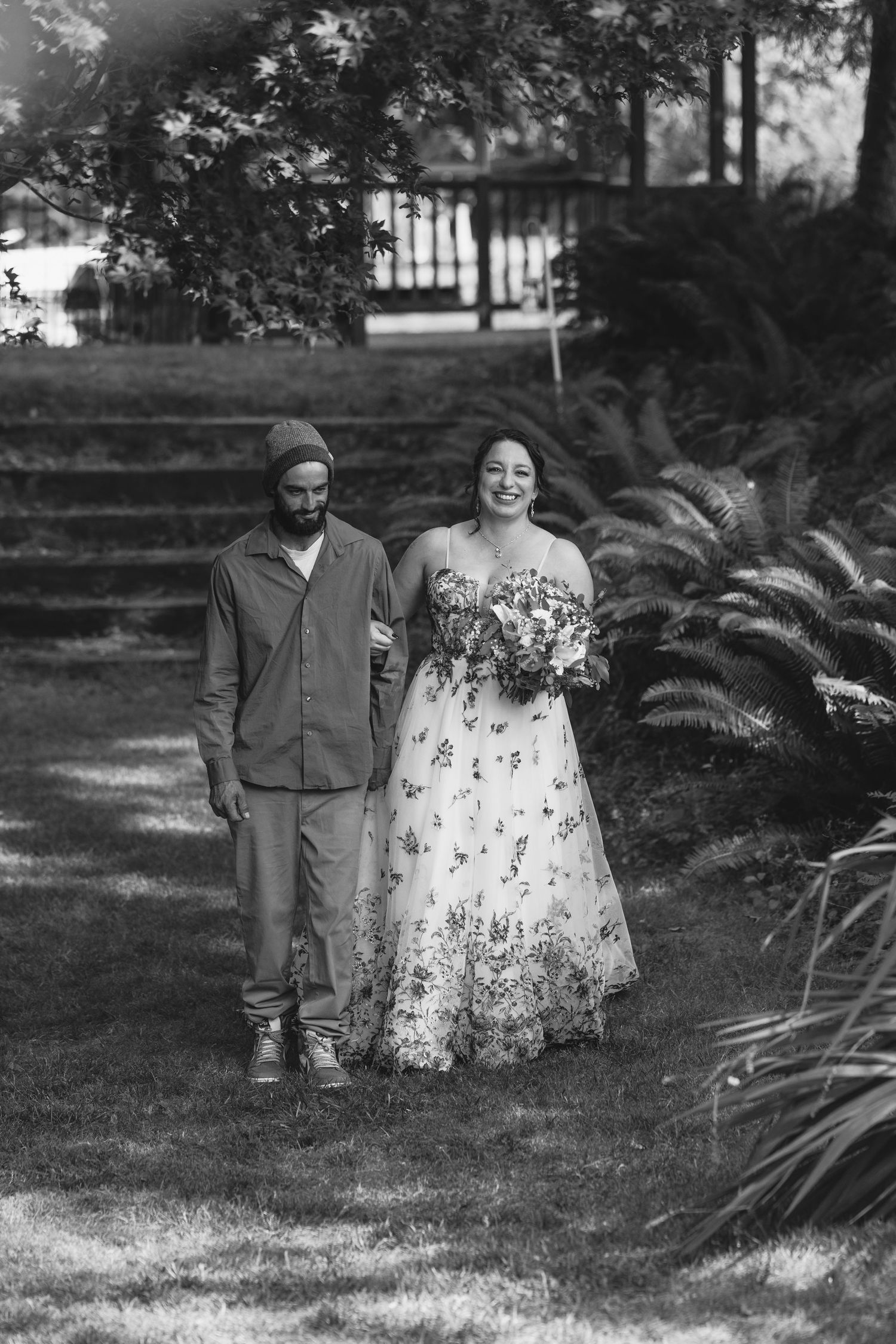 A couple stands together in a garden setting, with one person wearing a floral dress and the other in casual attire.