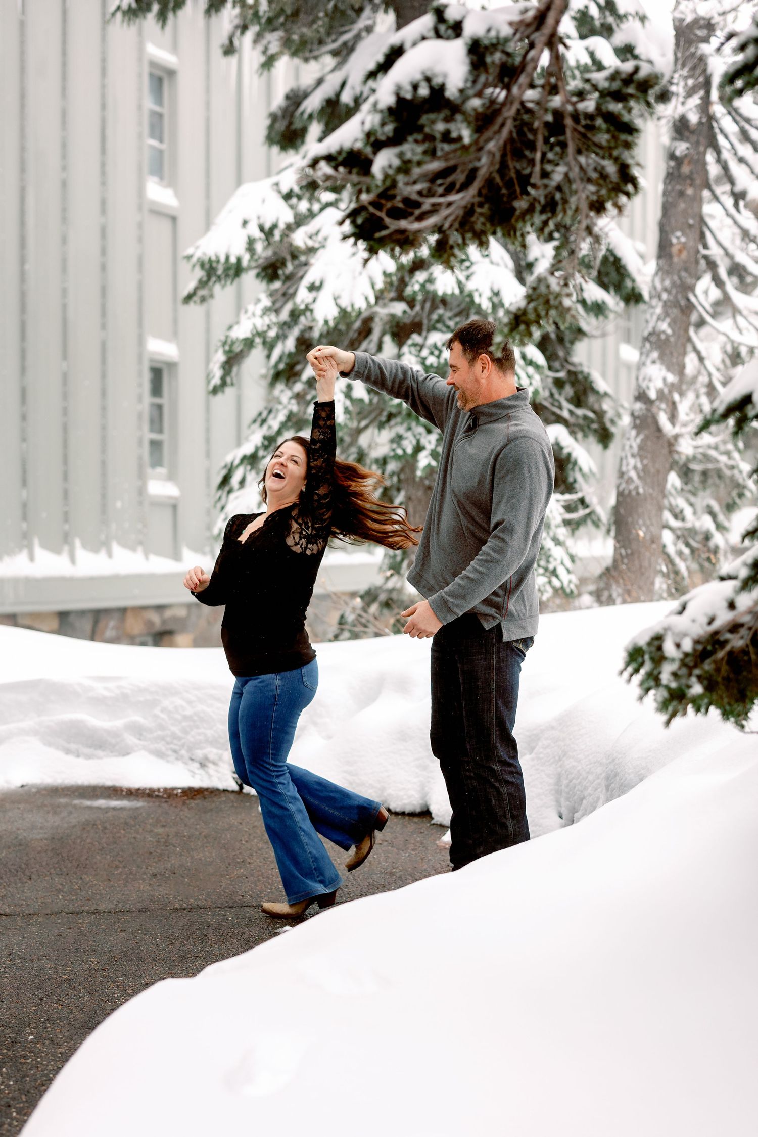 A couple plays together in a snowy winter landscape with evergreen trees and a building in the background.