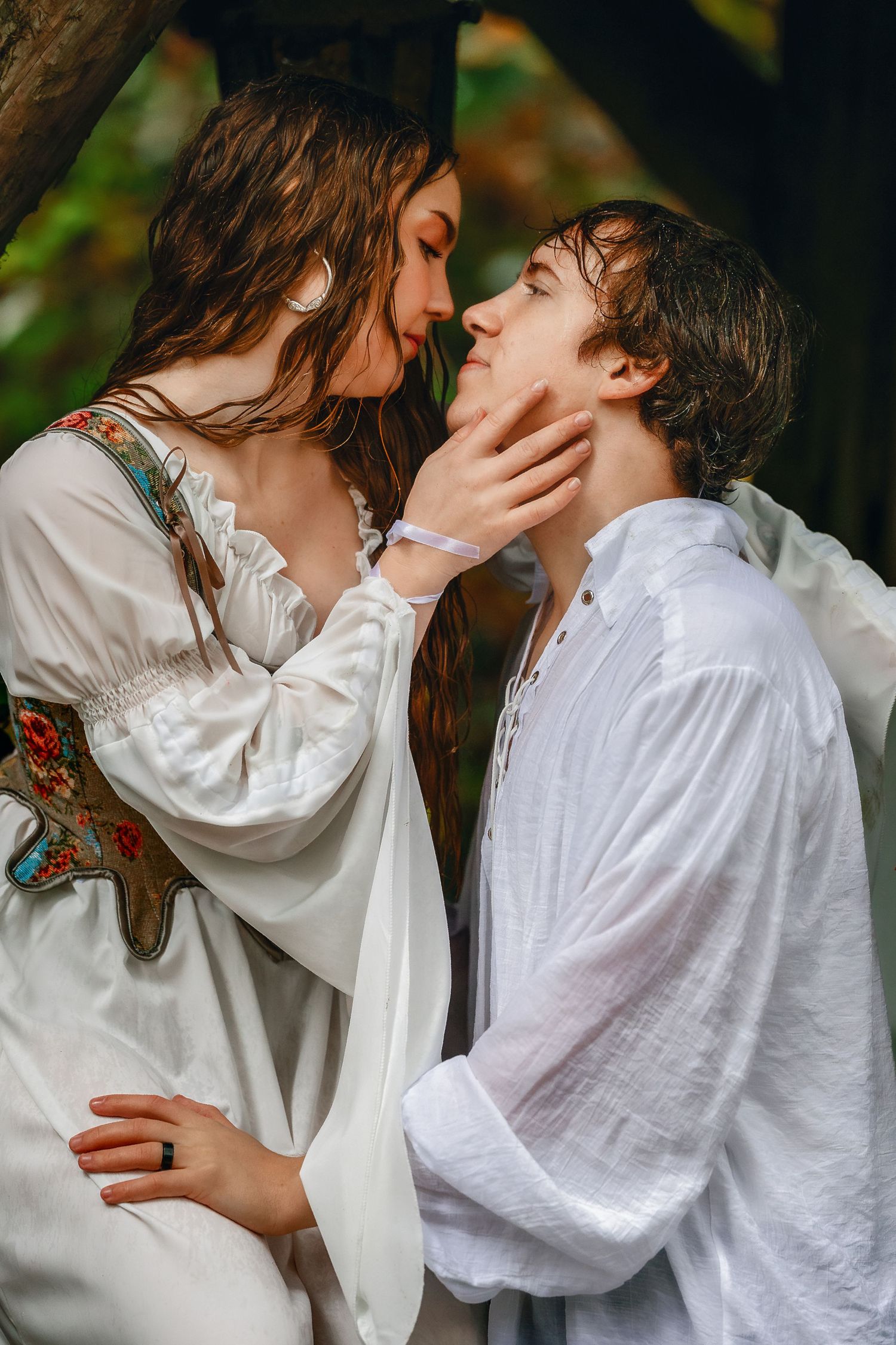 A romantic couple in white clothing share an intimate kiss outdoors against a natural backdrop.