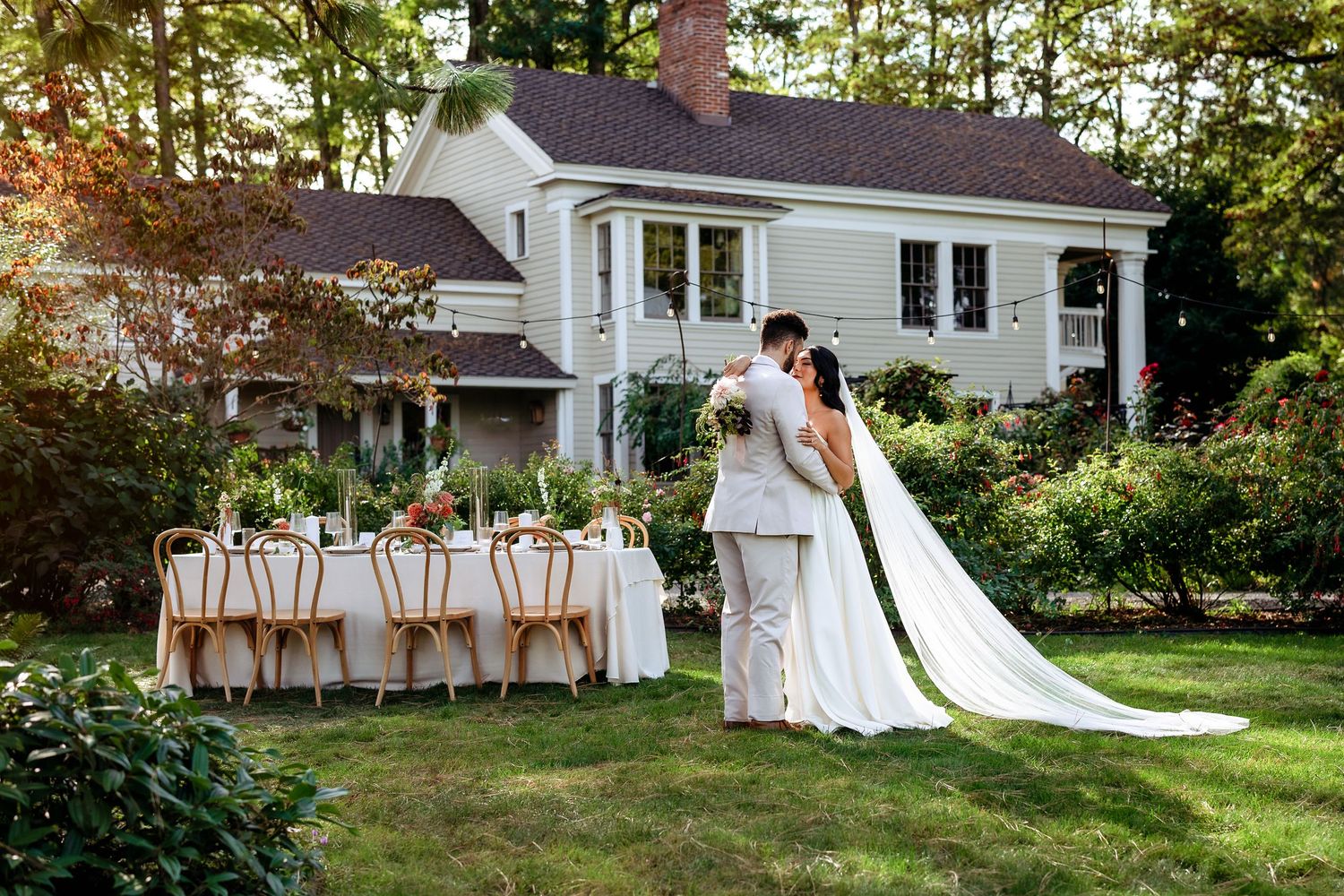 Elegant outdoor wedding reception setup with long table and white house in background during golden hour.