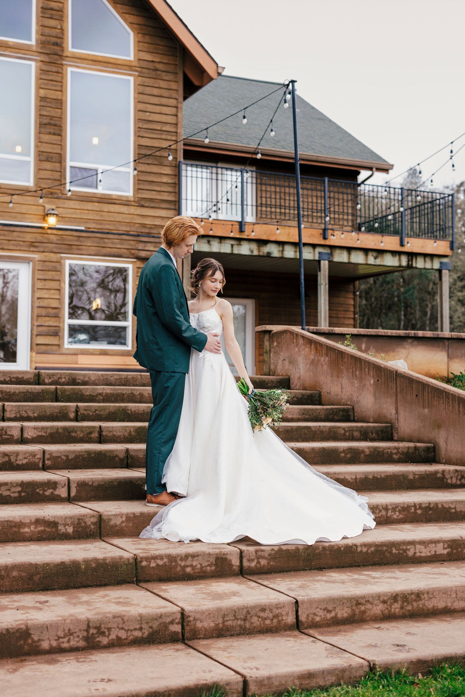 A couple stands together on stone steps outside a rustic wooden lodge during their wedding day.