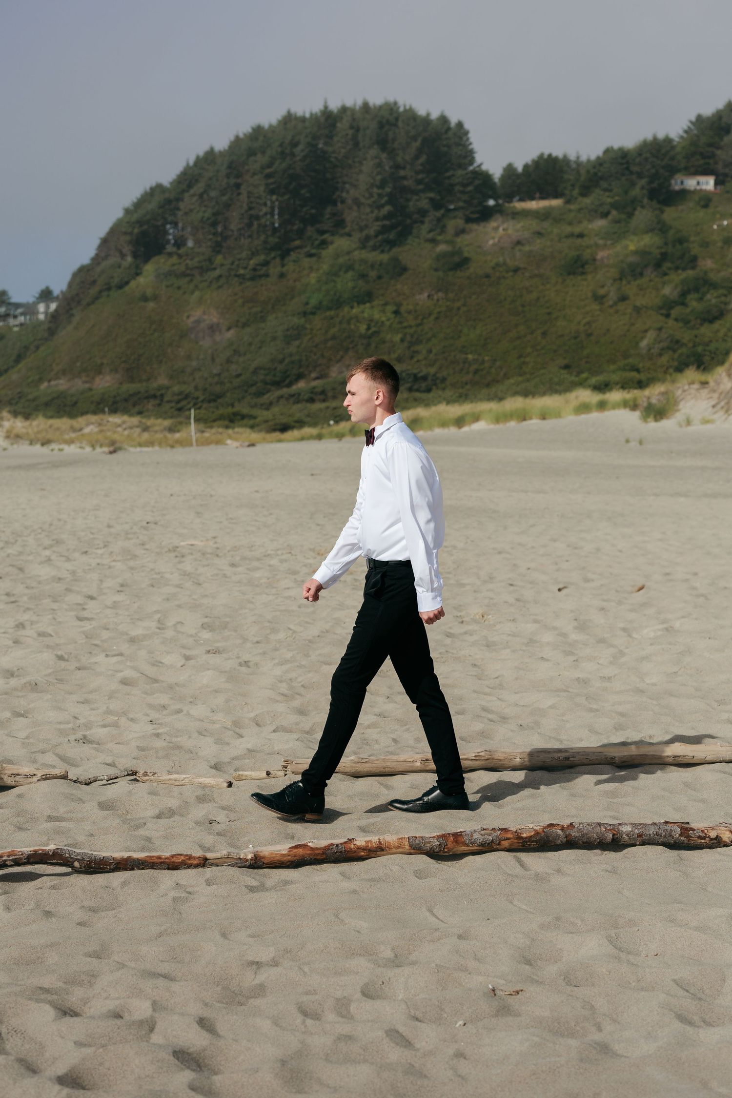 A person in formal attire walks along a scenic beach with forested cliffs in the background.