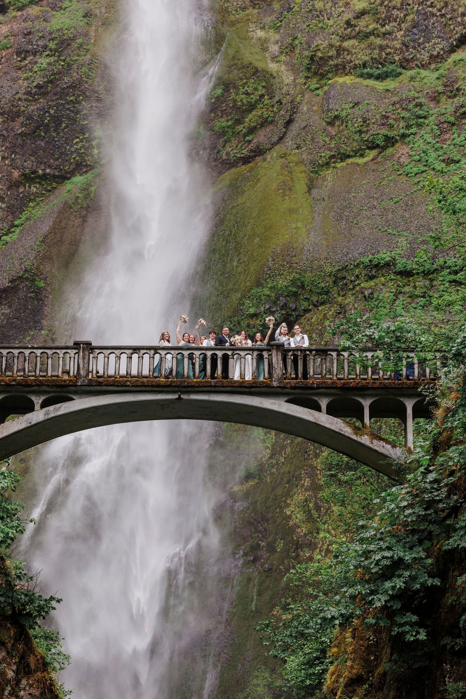 Wedding party gathered on scenic bridge overlooking dramatic Multnomah Falls waterfall in Oregon.