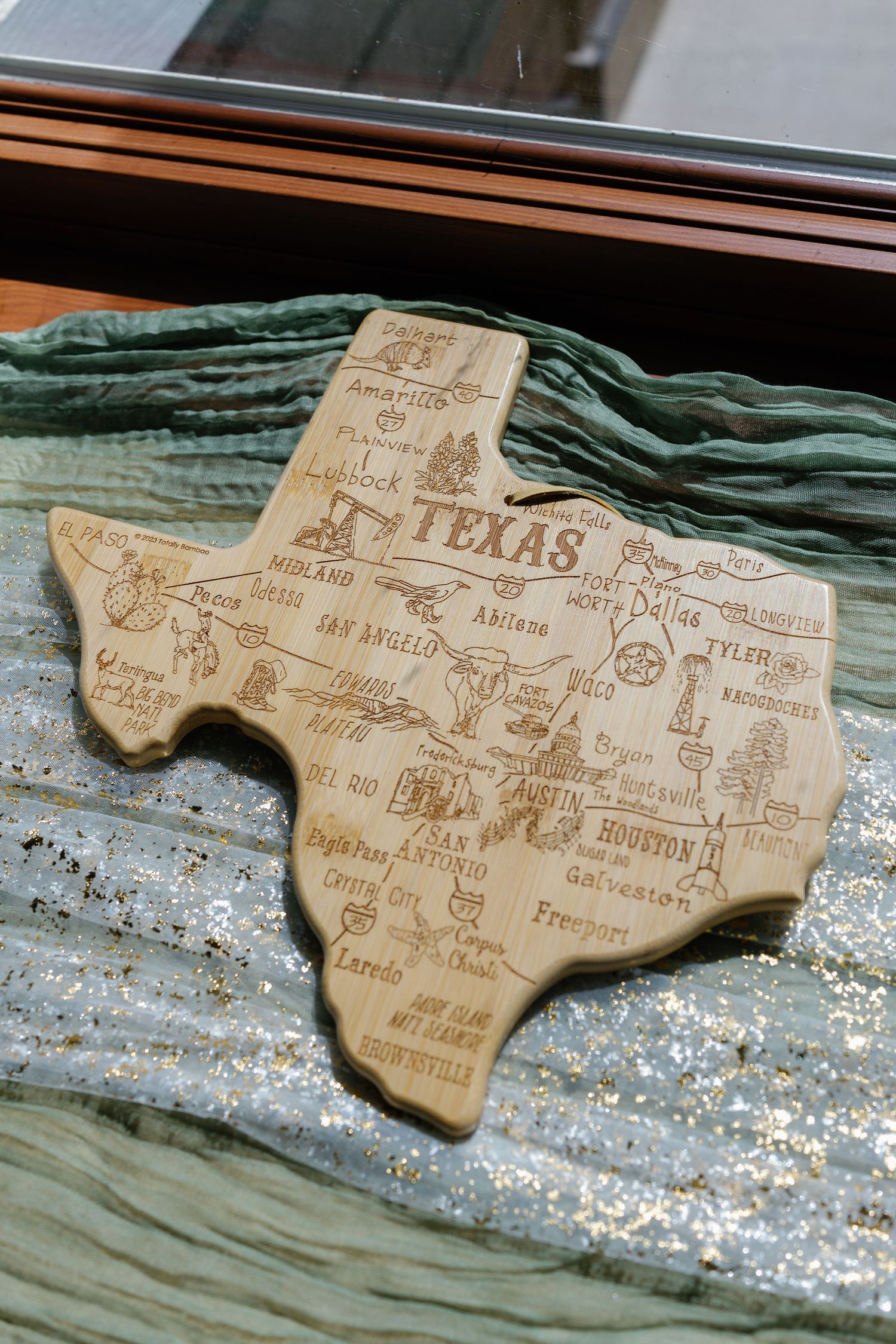 Wooden cutting board shaped like Texas with cities and landmarks engraved on a sparkly surface.