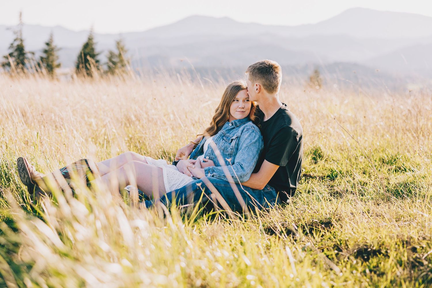 Couple sitting together in golden sunlit meadow with mountains and palm trees in background.