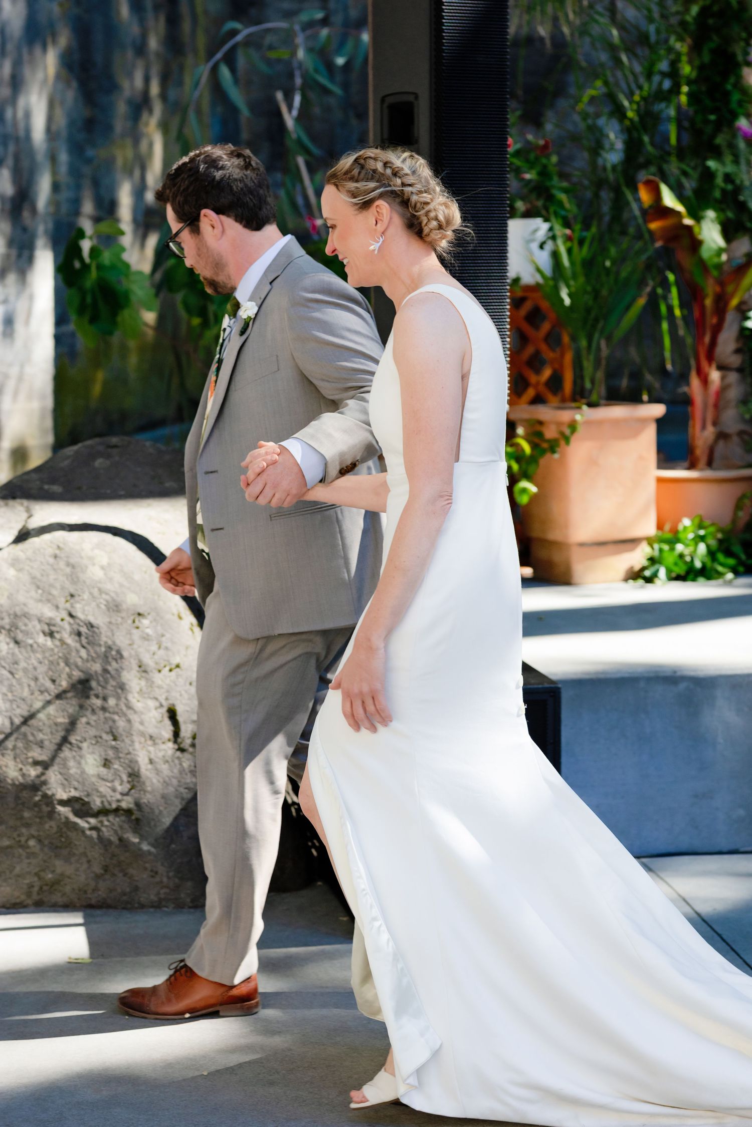 Bride in white gown and groom in gray suit walk hand in hand at outdoor wedding.