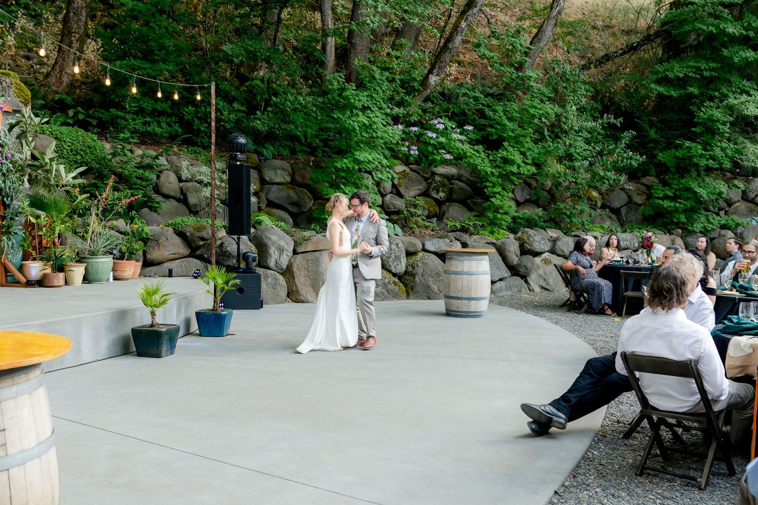 Newlyweds share romantic first dance on outdoor patio surrounded by wine barrels and seated wedding guests at dusk.