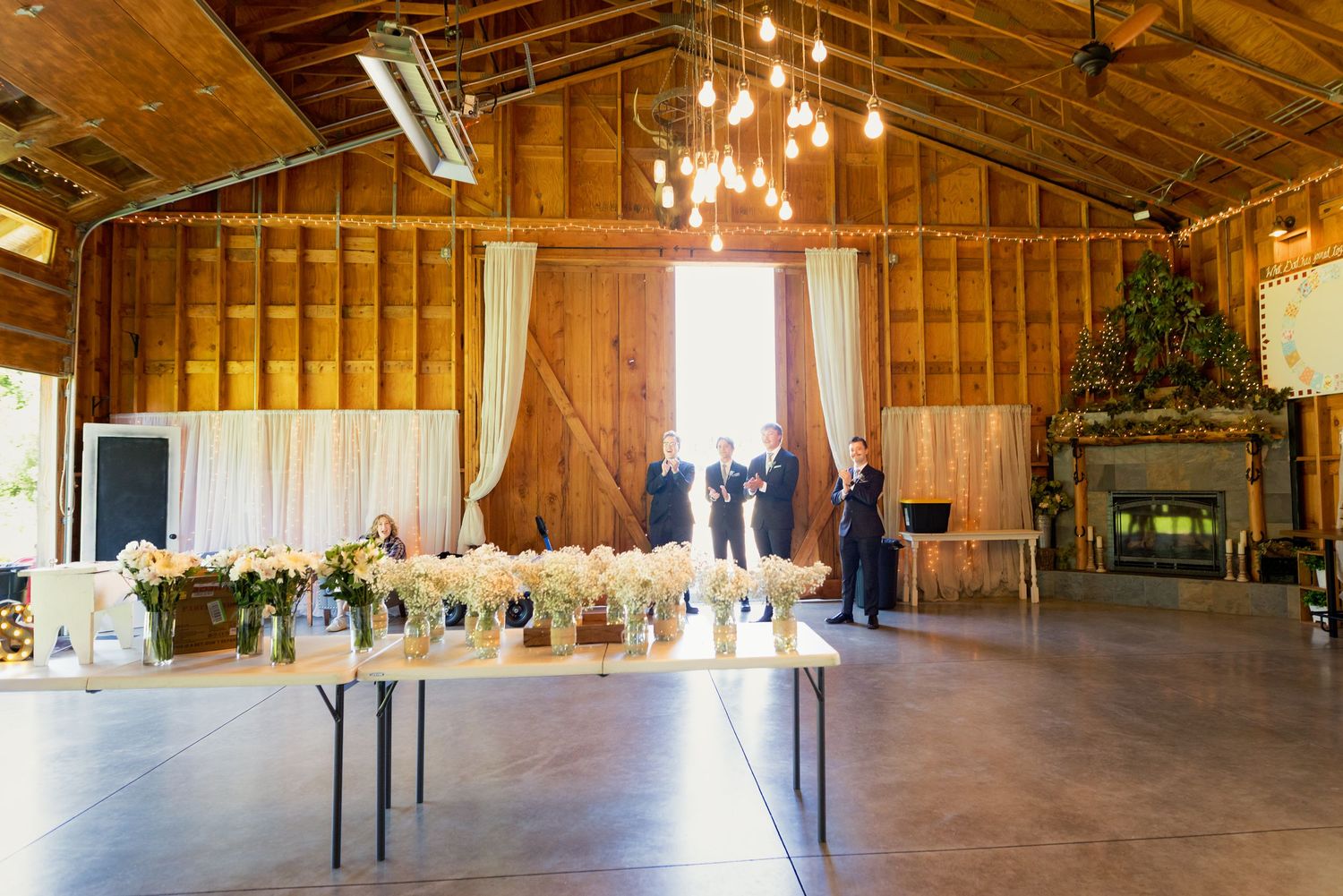 Rustic barn wedding venue with wooden walls, elegant chandelier, floral centerpieces on tables, and guests near a fireplace.