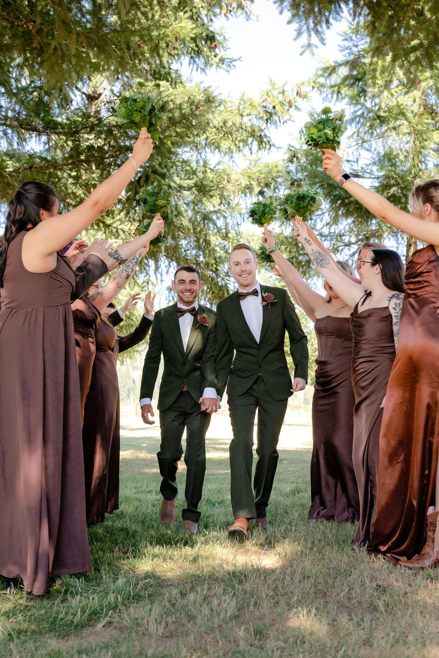 Two grooms in green tuxedos walk hand-in-hand through an arch of raised bouquets held by bridesmaids in brown gowns.