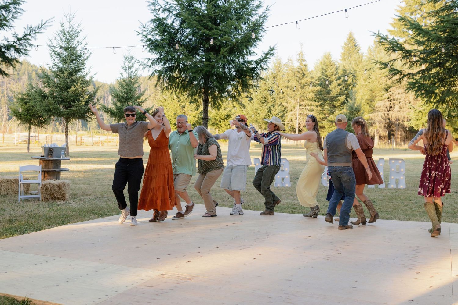 Wedding guests doing a conga line dance on outdoor patio surrounded by trees and string lights.