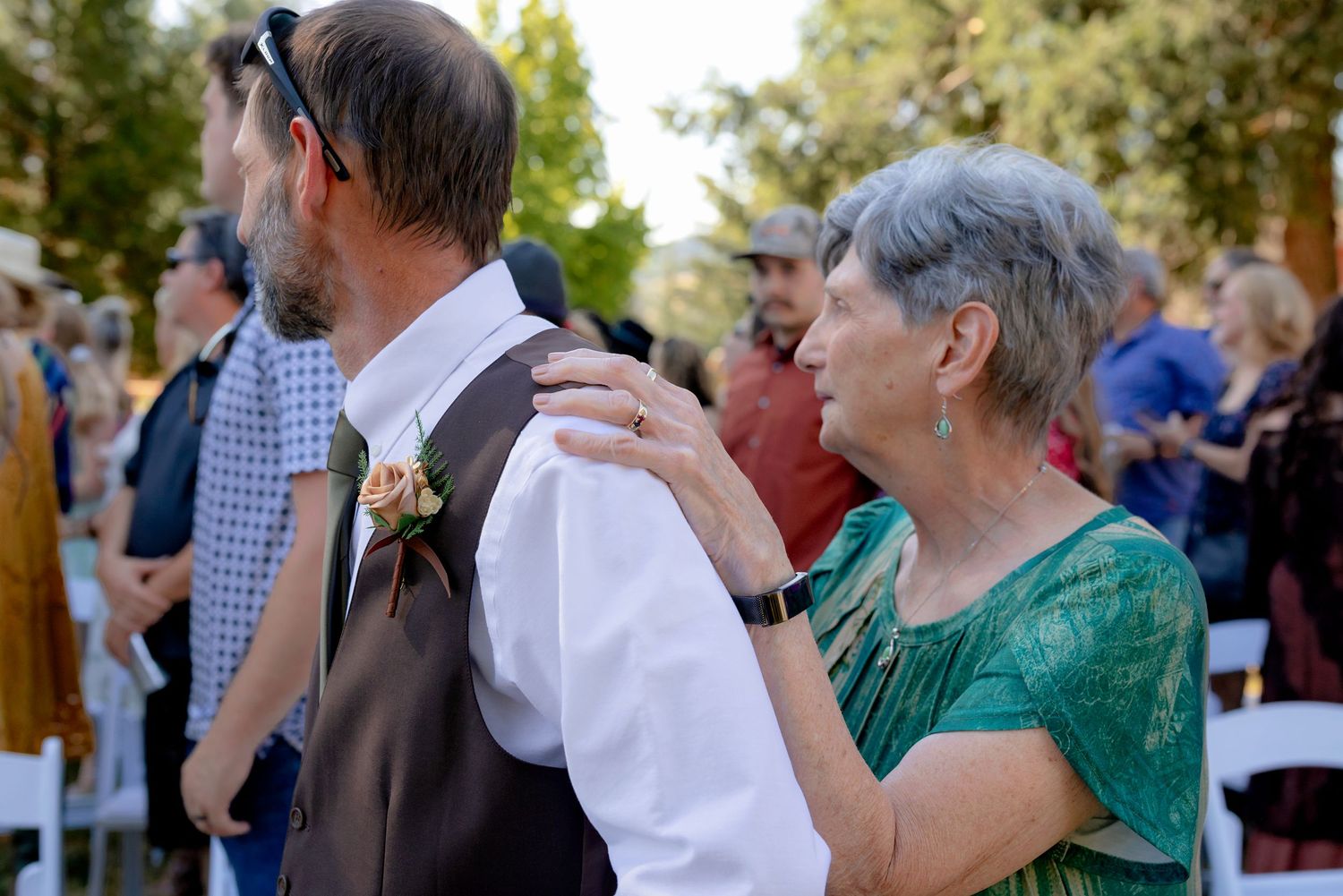 An elderly woman in green lace places her hand on a man in a brown vest with boutonniere at a wedding.