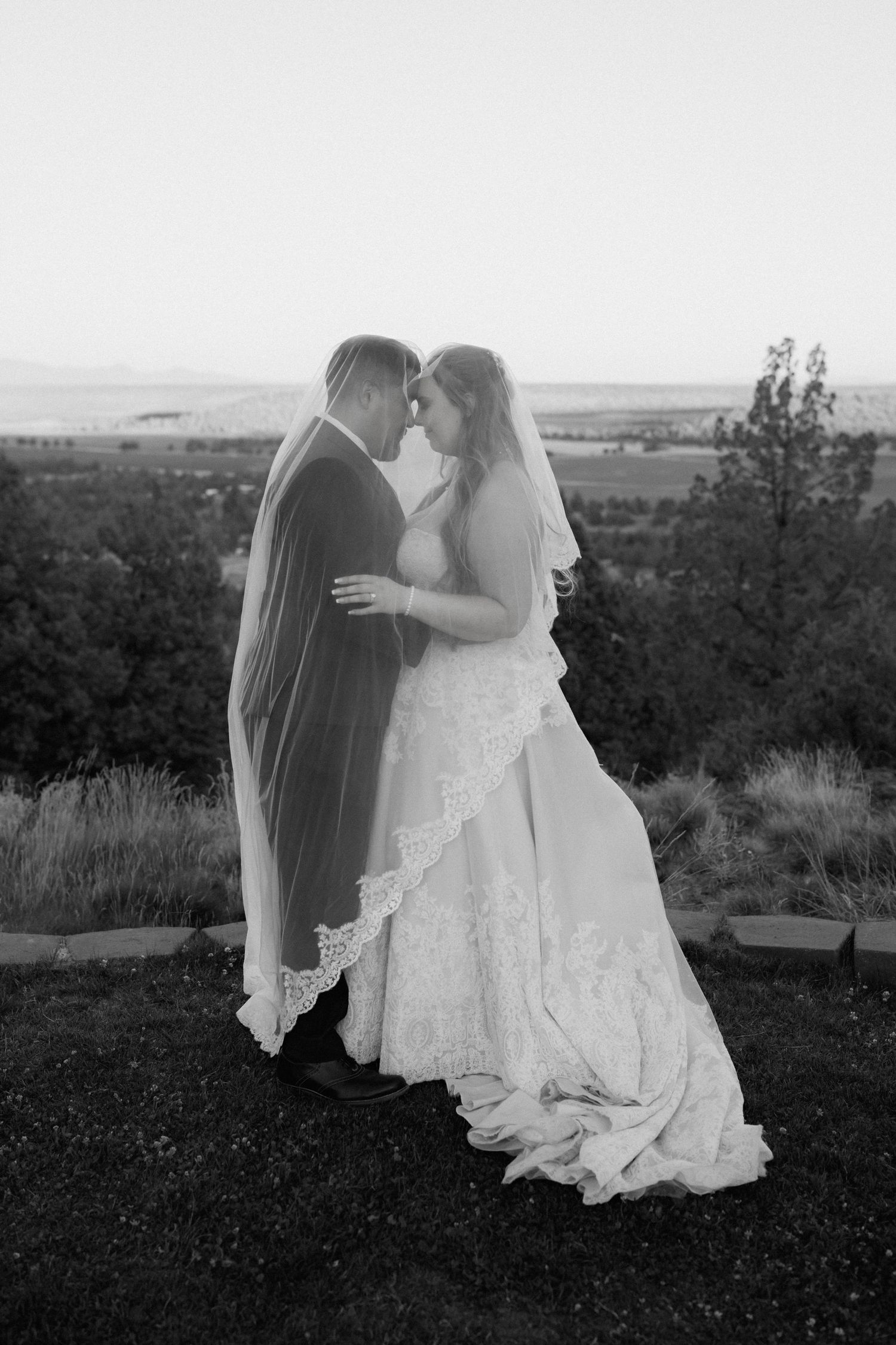 Black and white portrait of bride and groom touching foreheads under veil with scenic landscape behind.