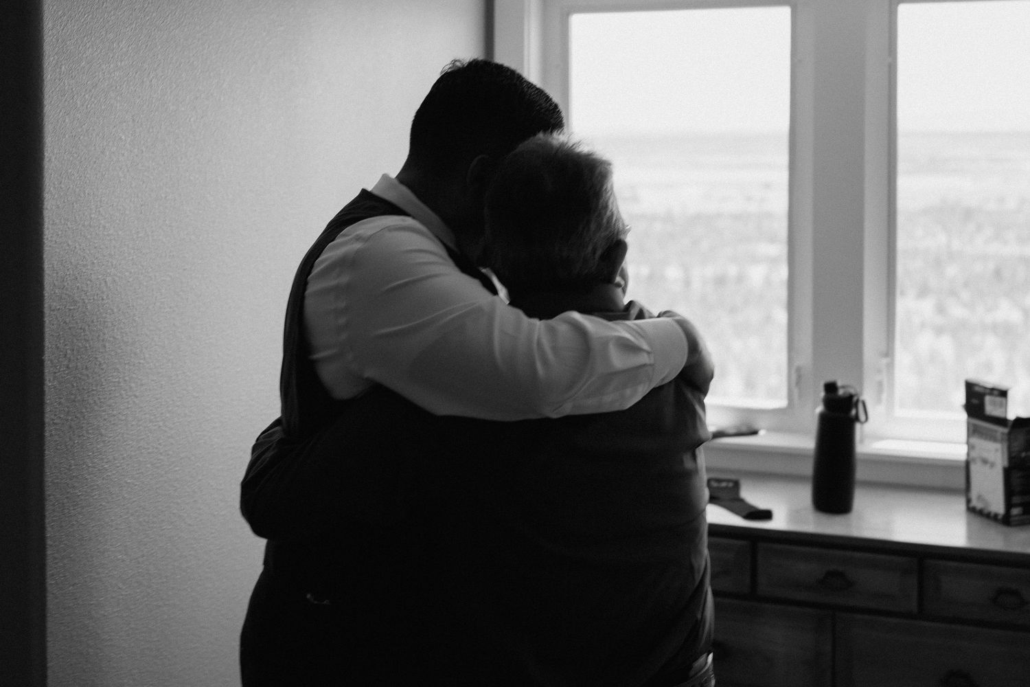 Groom and family member share an emotional embrace by a window in a black and white wedding photo.