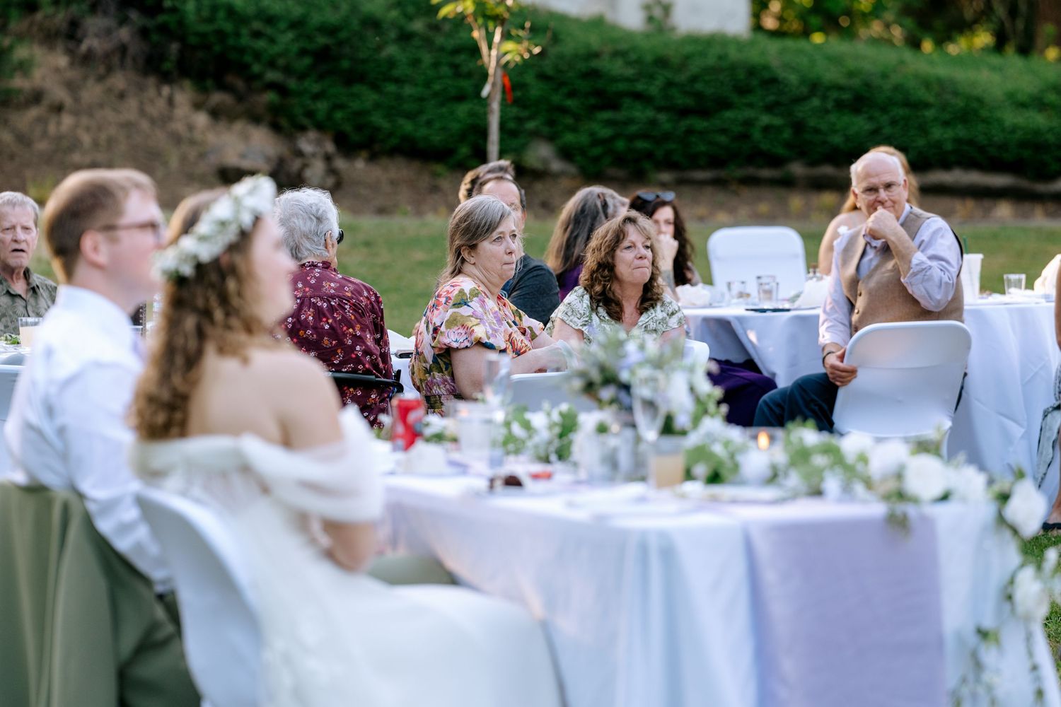 Bride wearing a floral crown seated at a decorated outdoor reception table surrounded by smiling wedding guests at dusk.